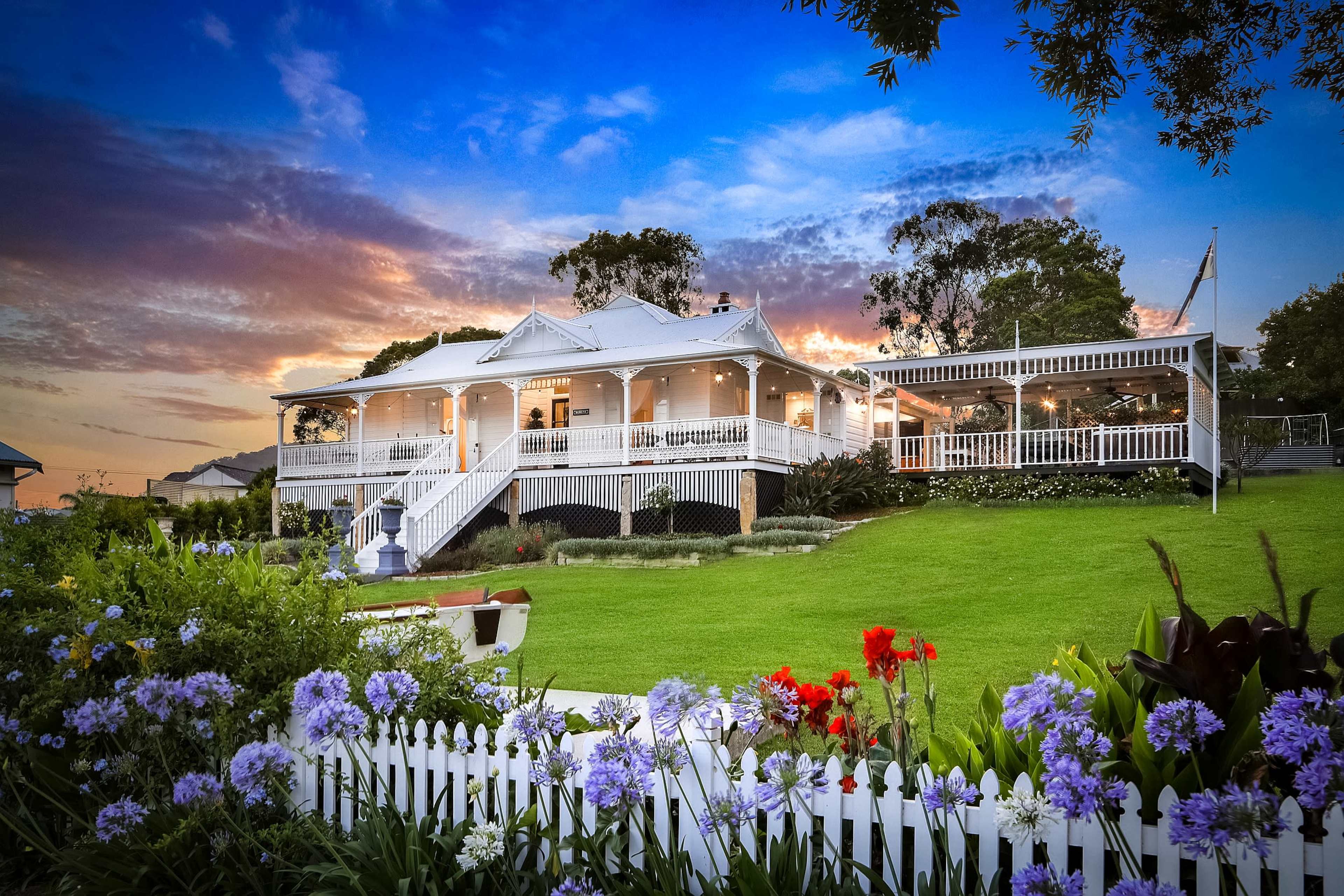 A large, two-story house with a wraparound veranda is set against a colorful sunset, surrounded by well-maintained gardens and vibrant flowers.