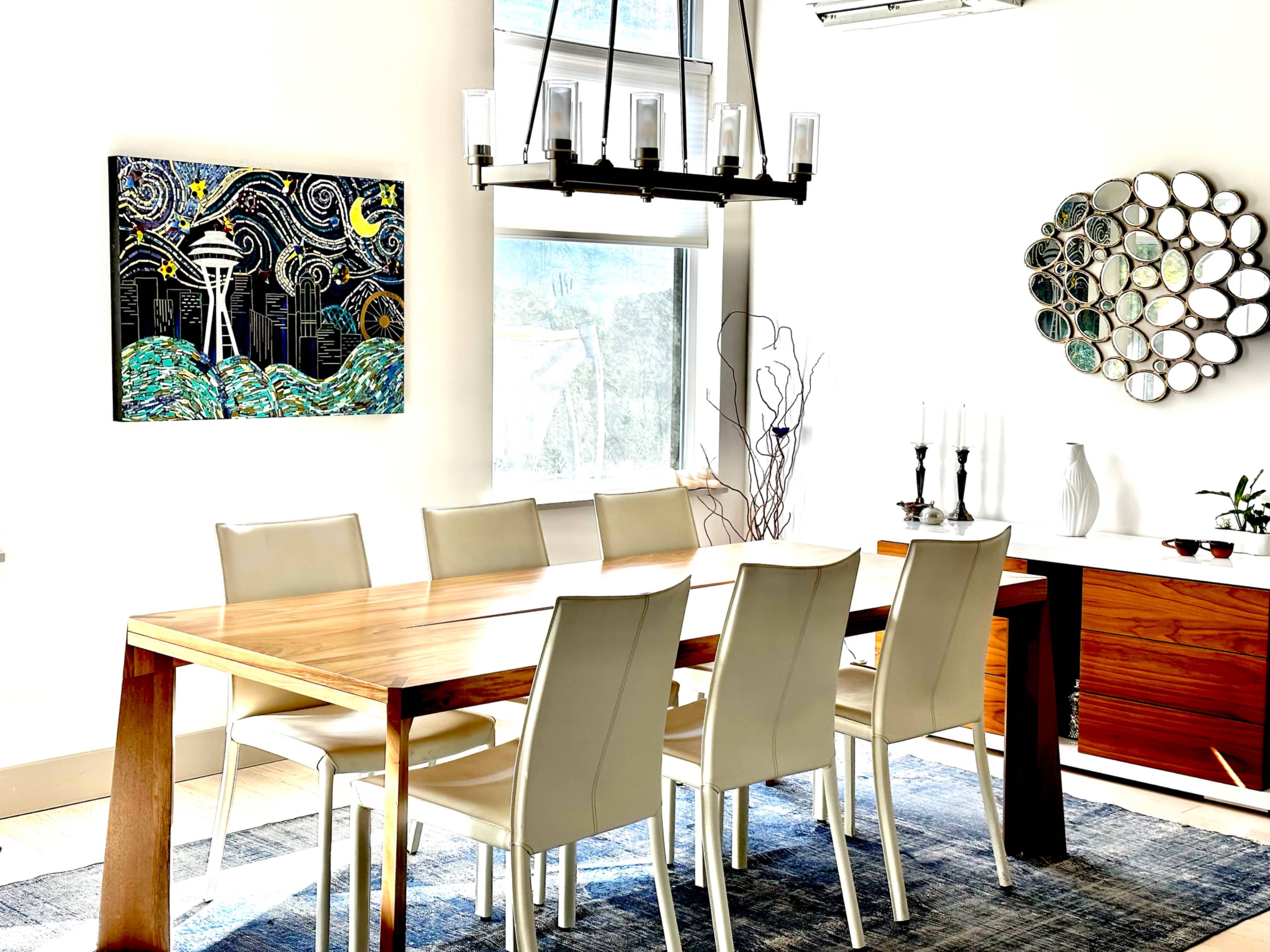 A modern dining room features a long wooden table surrounded by white chairs, with artwork on the wall and decorative pieces nearby.