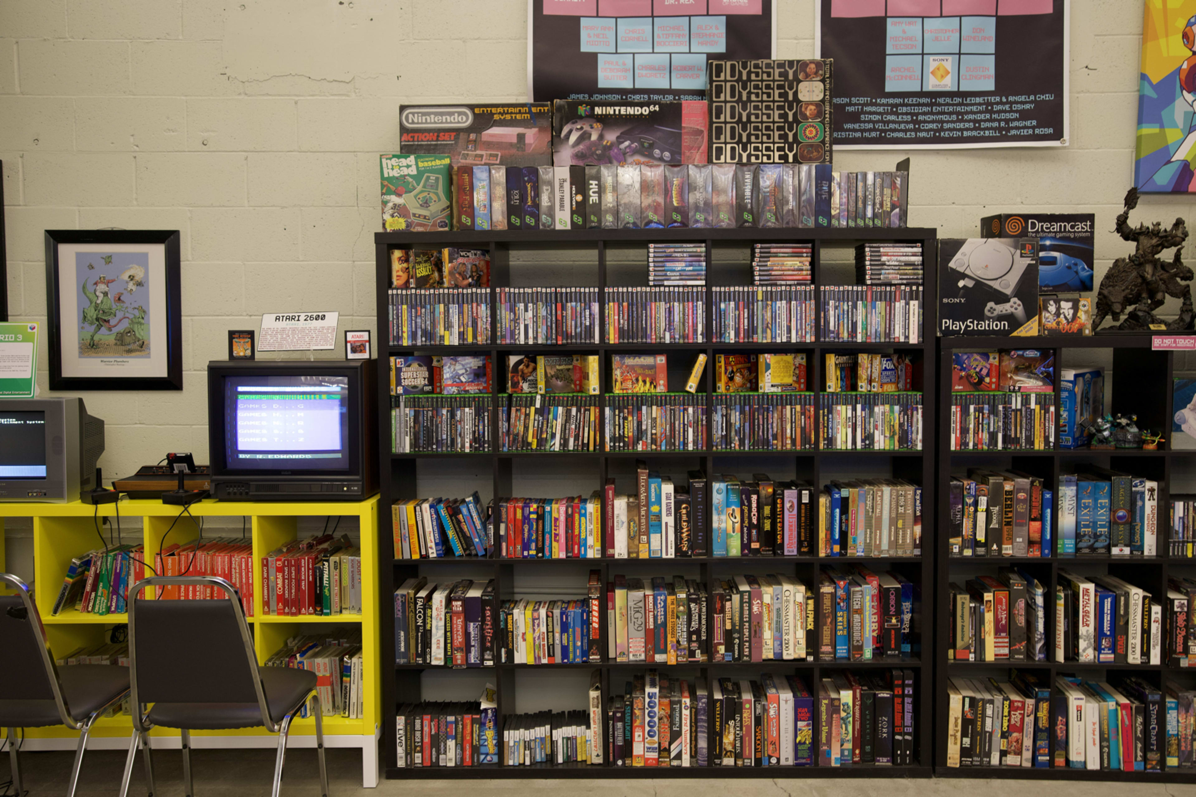 A well-organized display of video games and consoles on shelves, with a retro TV and chairs in the foreground.