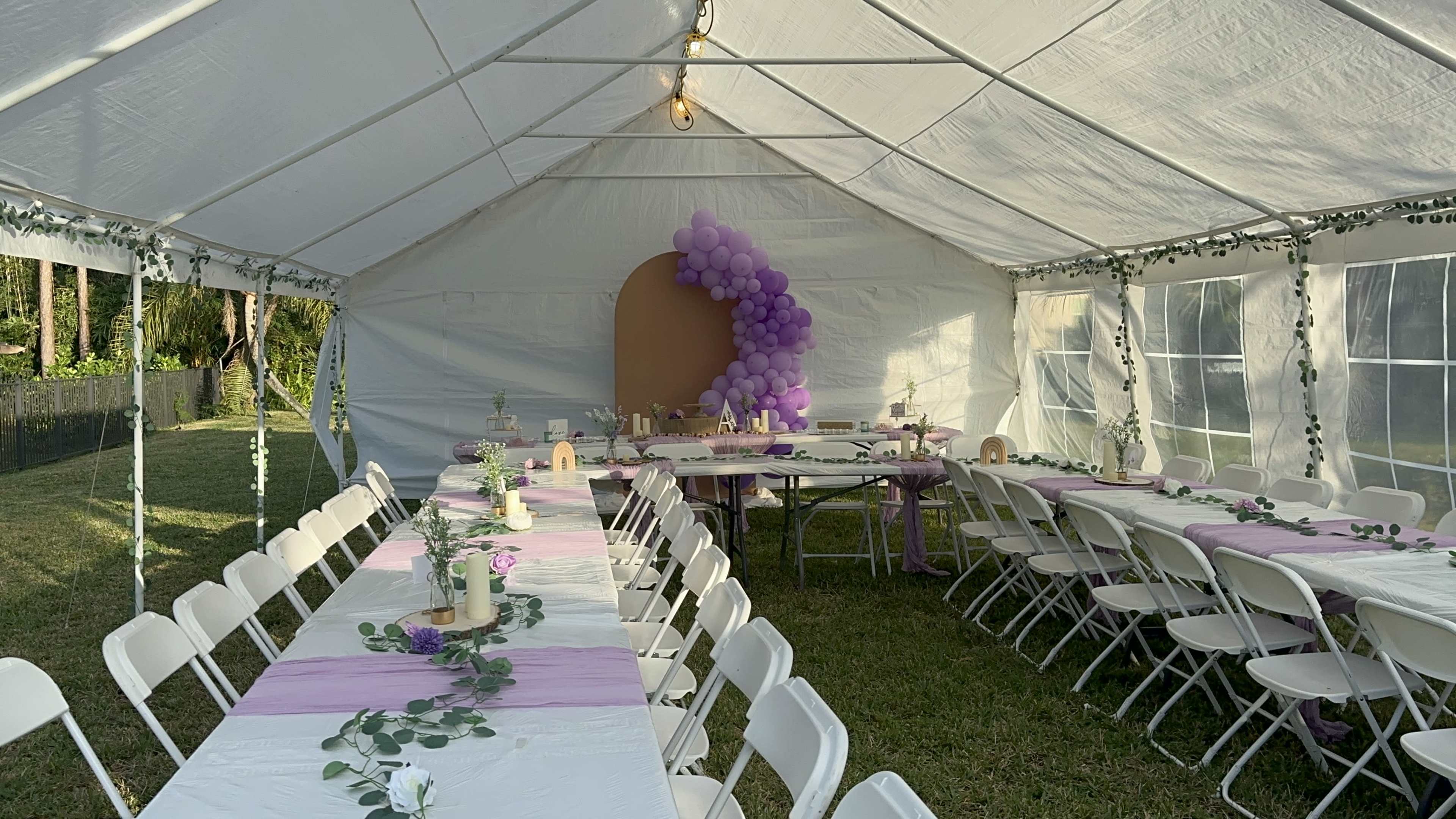 A tent is set up for a party, featuring long tables arranged with white tablecloths, lavender accents, and decorations, along with a balloon arch in the corner.