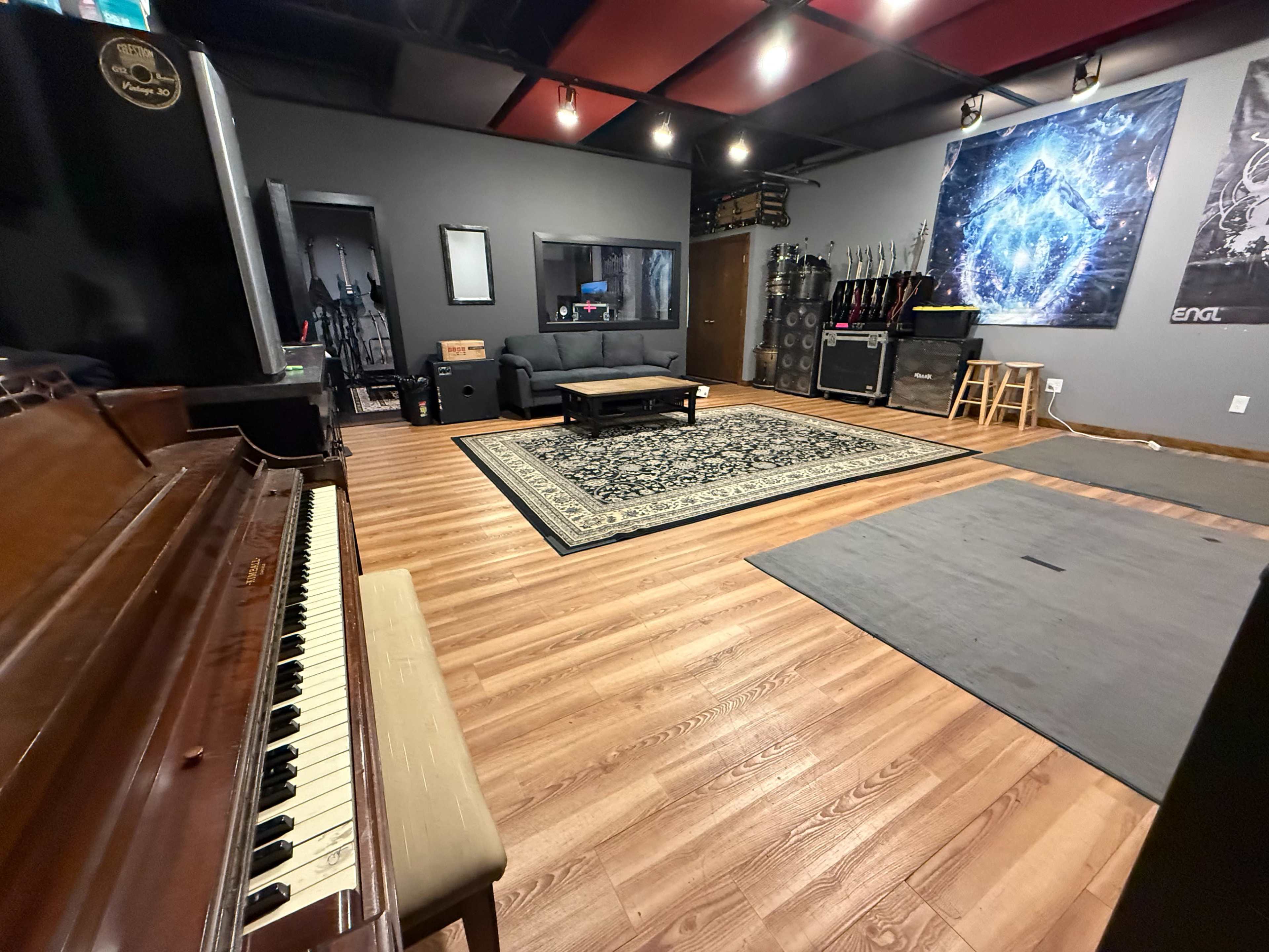 The image shows a spacious music studio with wooden floors, a grand piano in the foreground, and various musical instruments and equipment displayed along the walls.