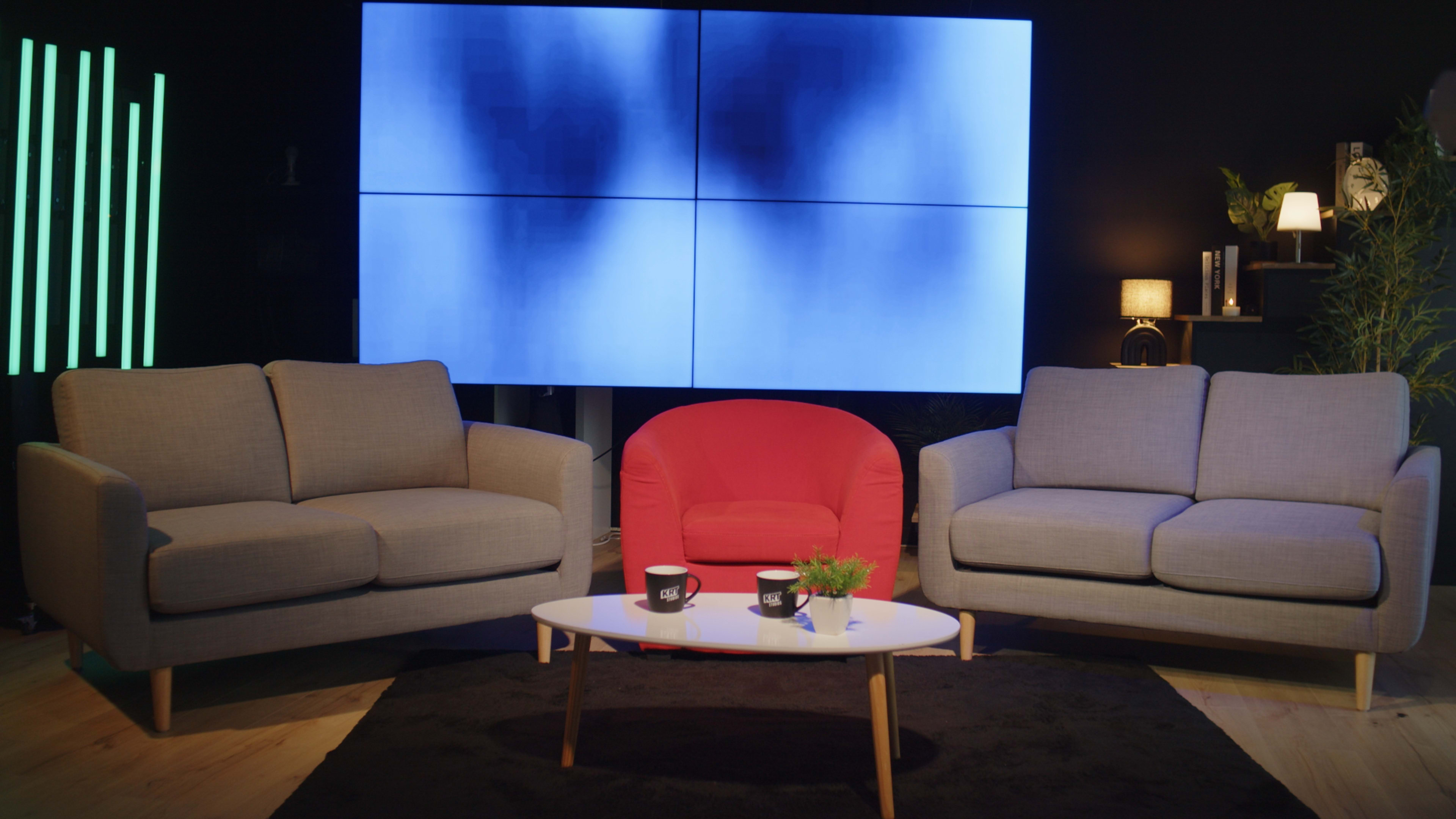The image shows a modern interior setup featuring two gray sofas flanking a bright red armchair, arranged around a white coffee table on a black rug in front of a large video wall.