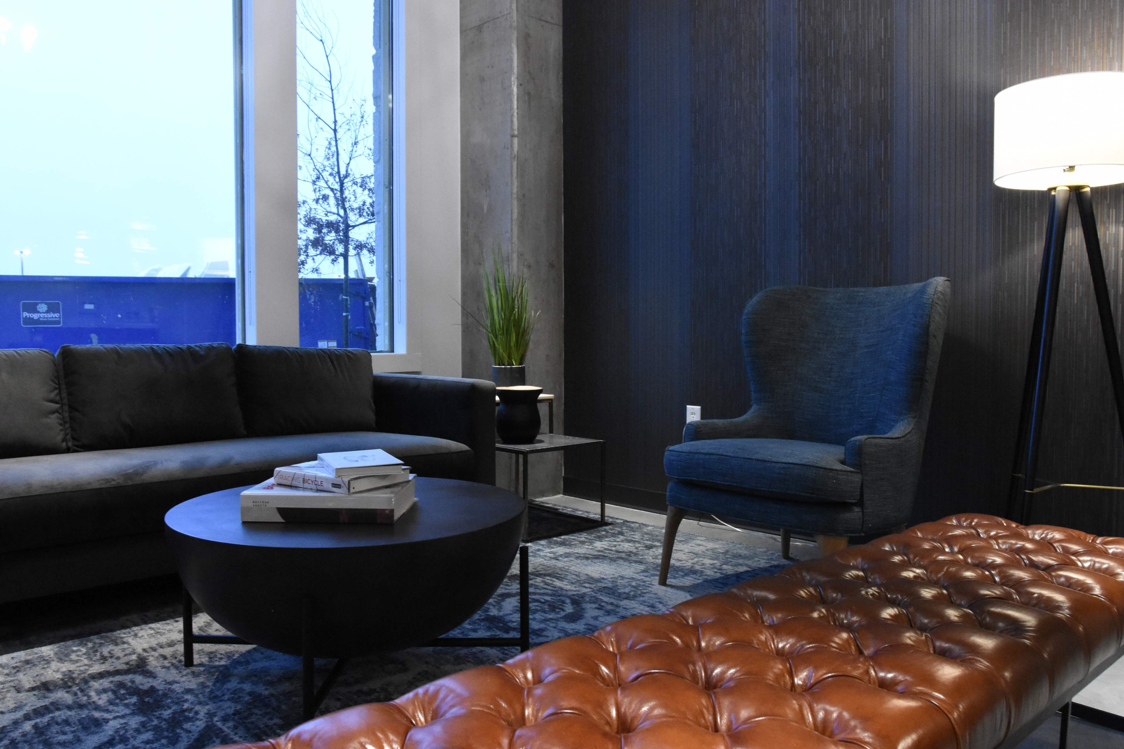 A modern living room features a gray sofa, a blue armchair, a round black coffee table, and a leather-tufted ottoman under soft lighting.