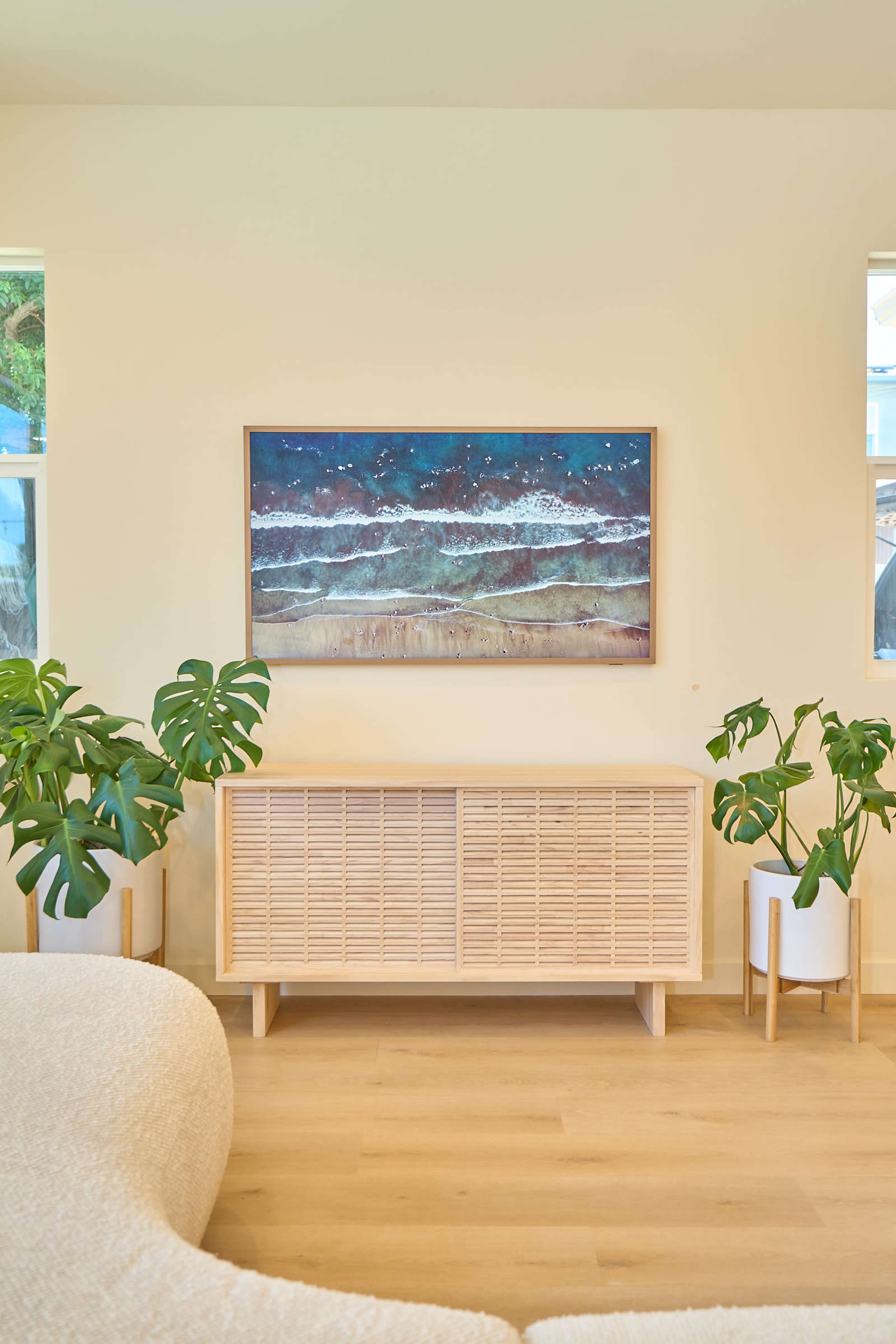 The scene features a light-colored room with a wooden sideboard, a piece of ocean-themed artwork on the wall, and two potted plants on either side of the sideboard.