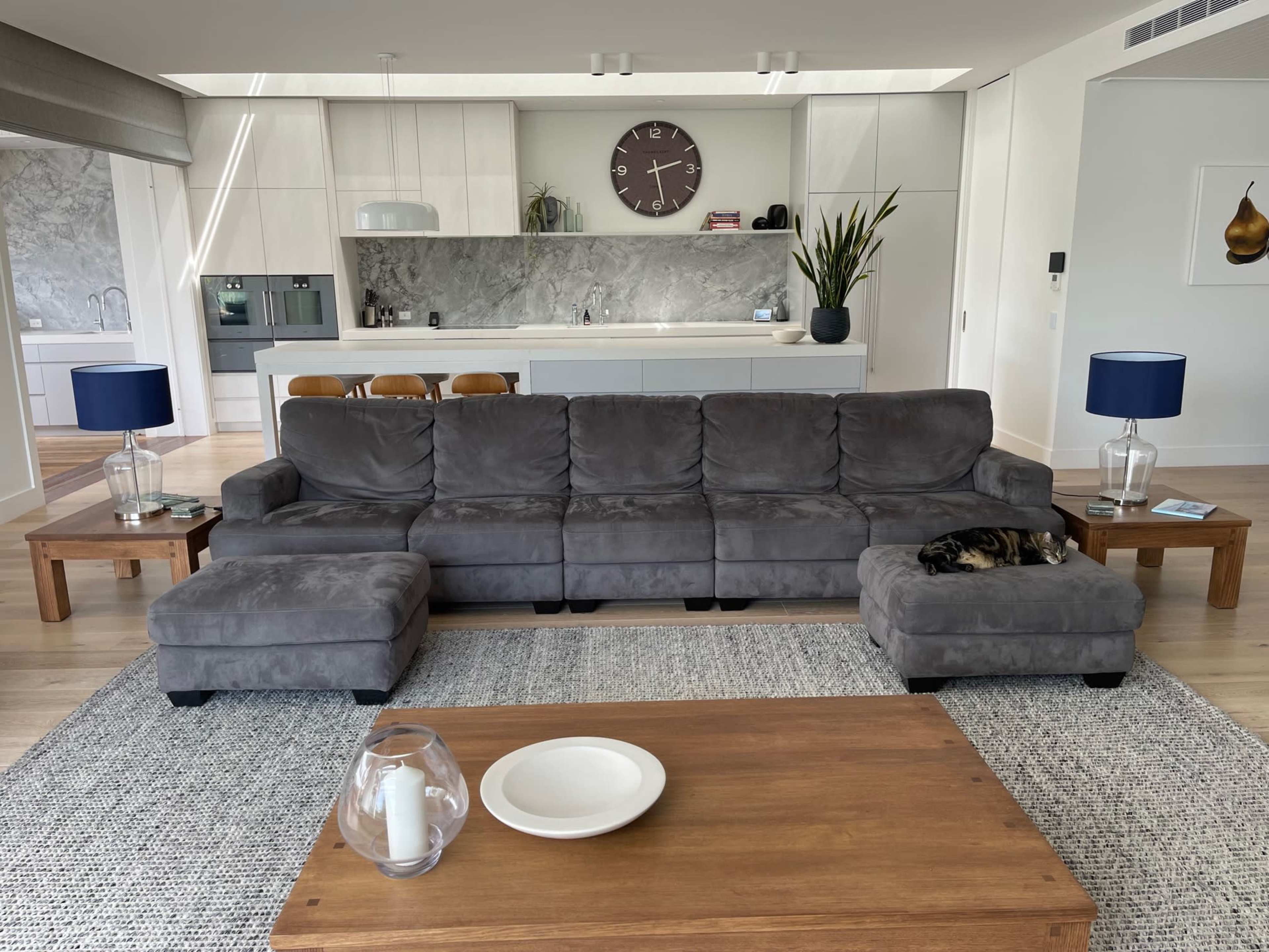 A gray sectional sofa with a matching ottoman is positioned in a bright living space featuring a wooden coffee table, two blue lamps, and a modern kitchen in the background.