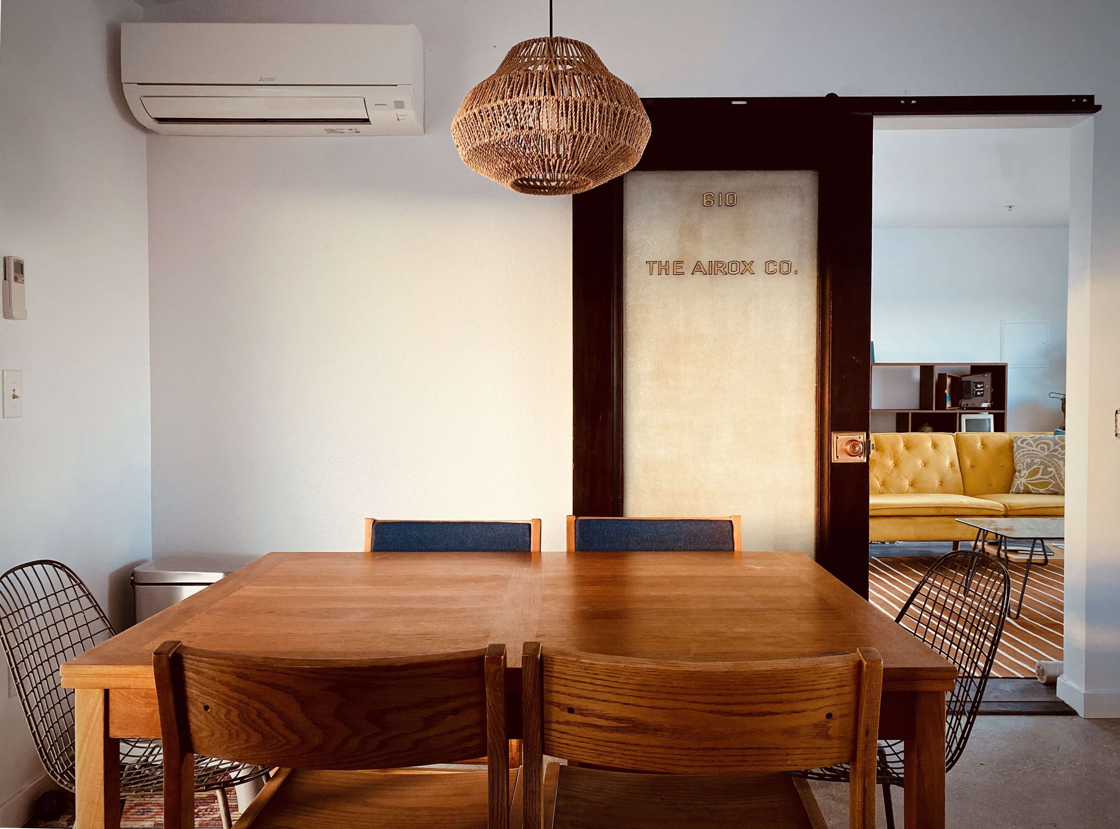 A wooden dining table with four chairs is positioned in front of a sliding door labeled "THE ARROW CO.", with a woven pendant light hanging above.