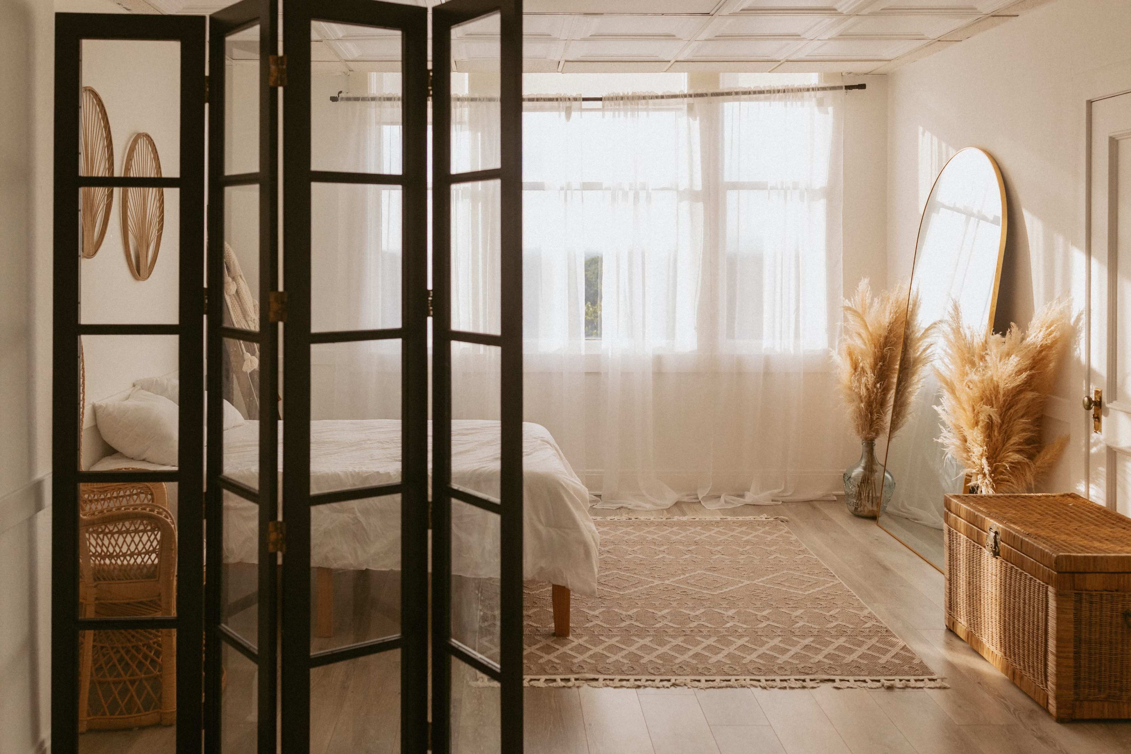 A bright and airy bedroom features a bed with white linens, a large mirror, natural decor elements, and a folding screen that provides separation from the rest of the space.