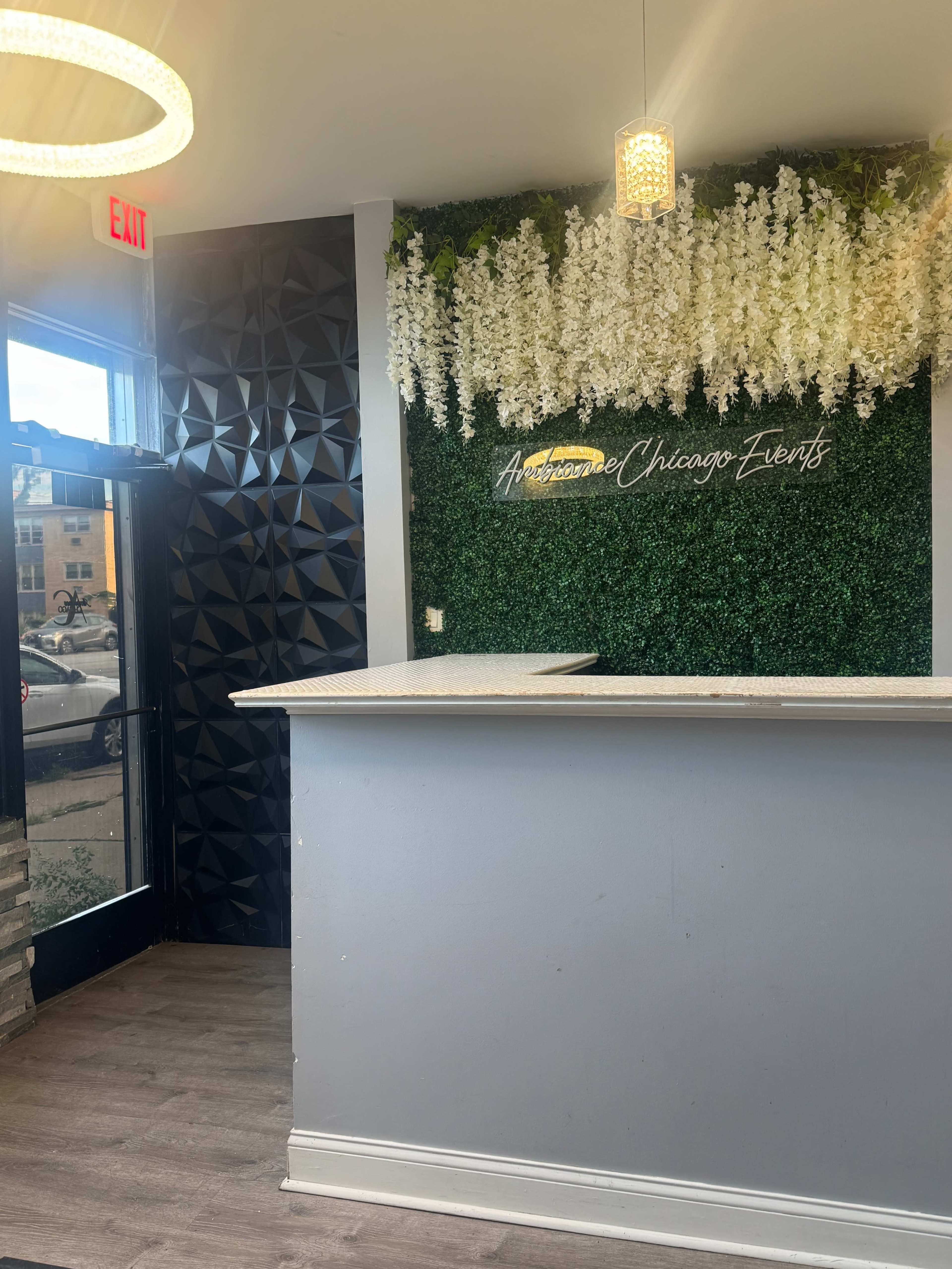 The image shows a reception area with a counter, a wall featuring greenery and hanging white flowers, and an exit door on the left.