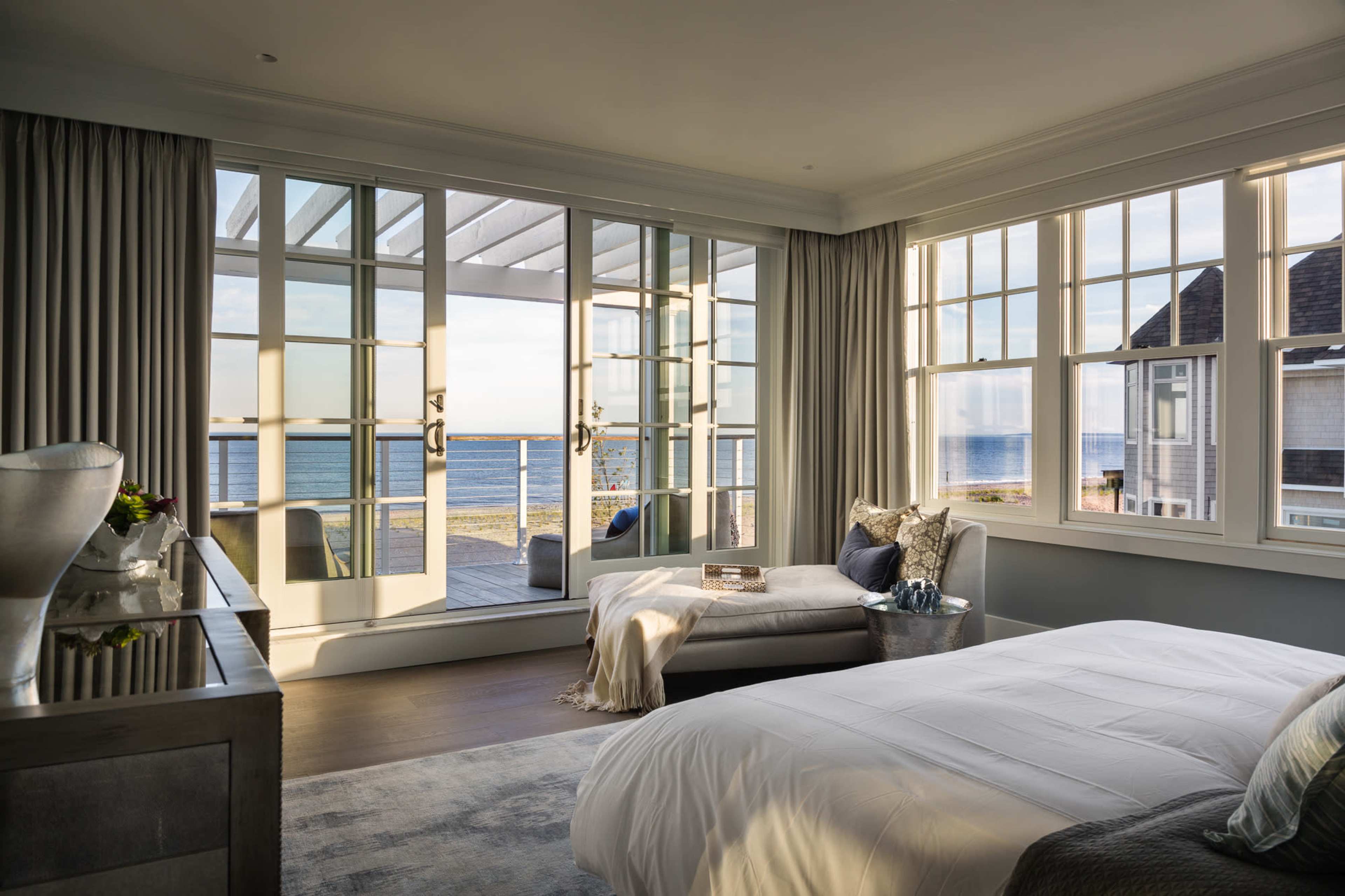 A spacious bedroom features large windows overlooking the ocean, with a seating area and a bed dressed in white linens.