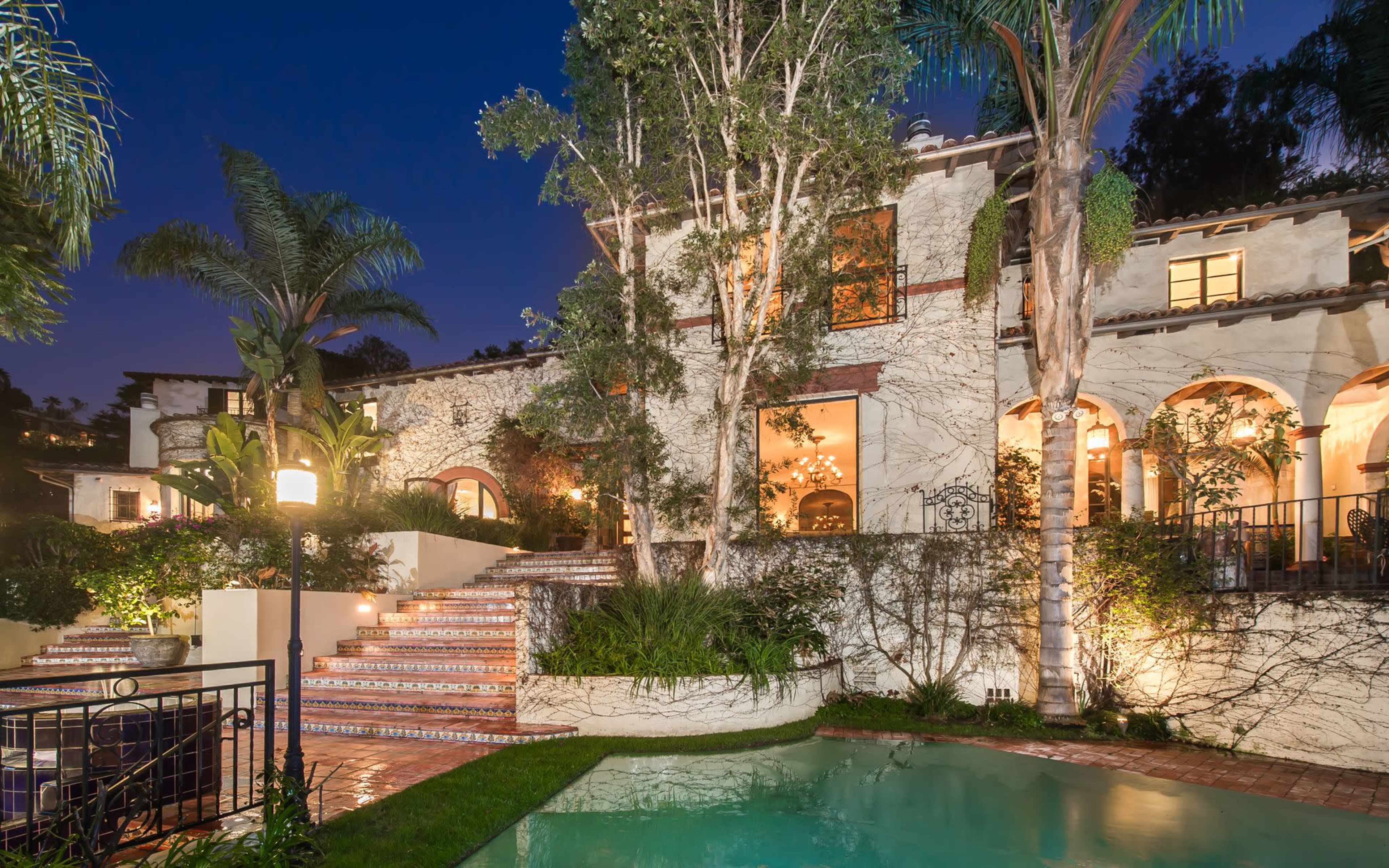 A large, elegantly designed home features arched windows and a pool surrounded by lush landscaping, illuminated by soft outdoor lighting.
