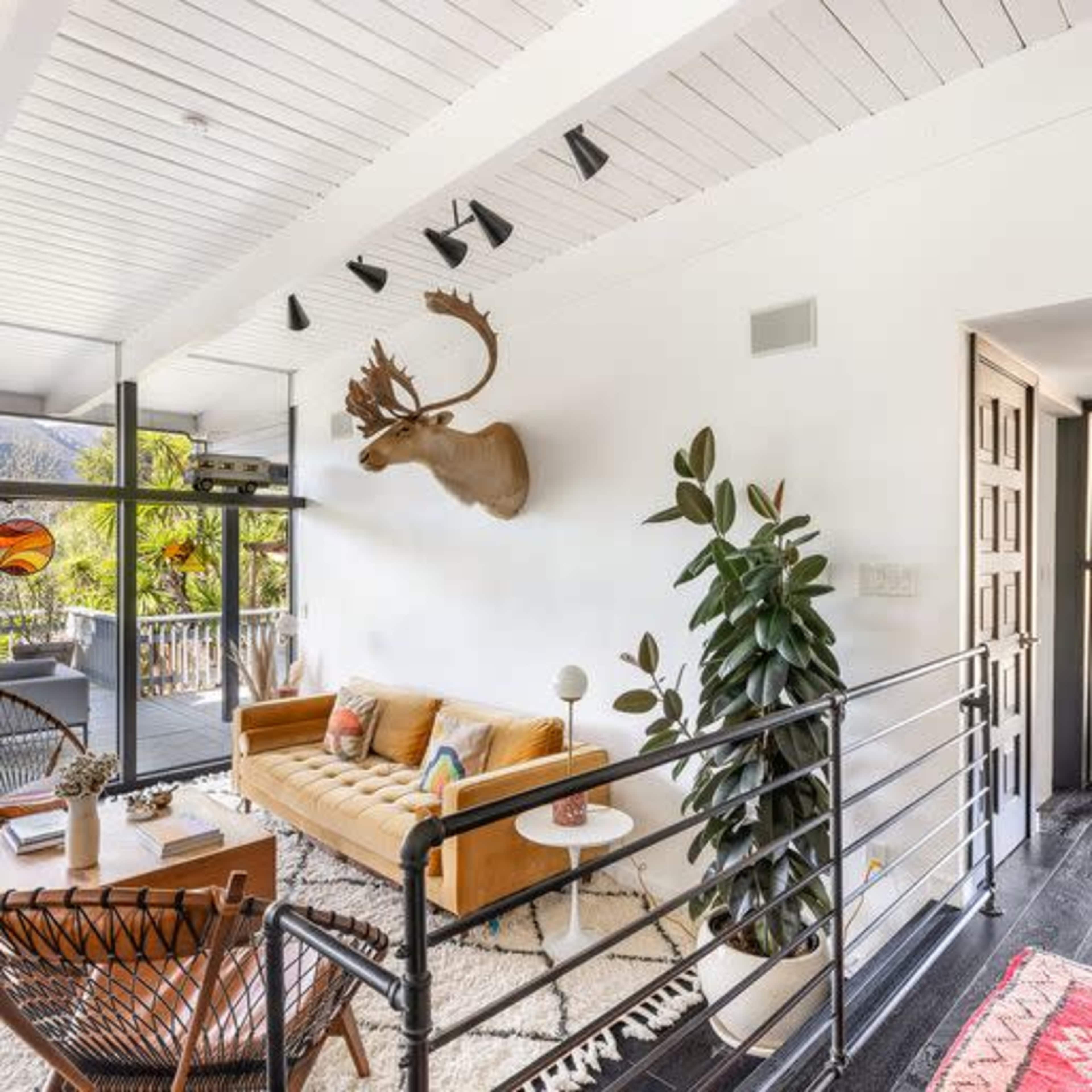 A modern living room with a yellow couch, a mounted deer head on the wall, and a staircase leading to another room.