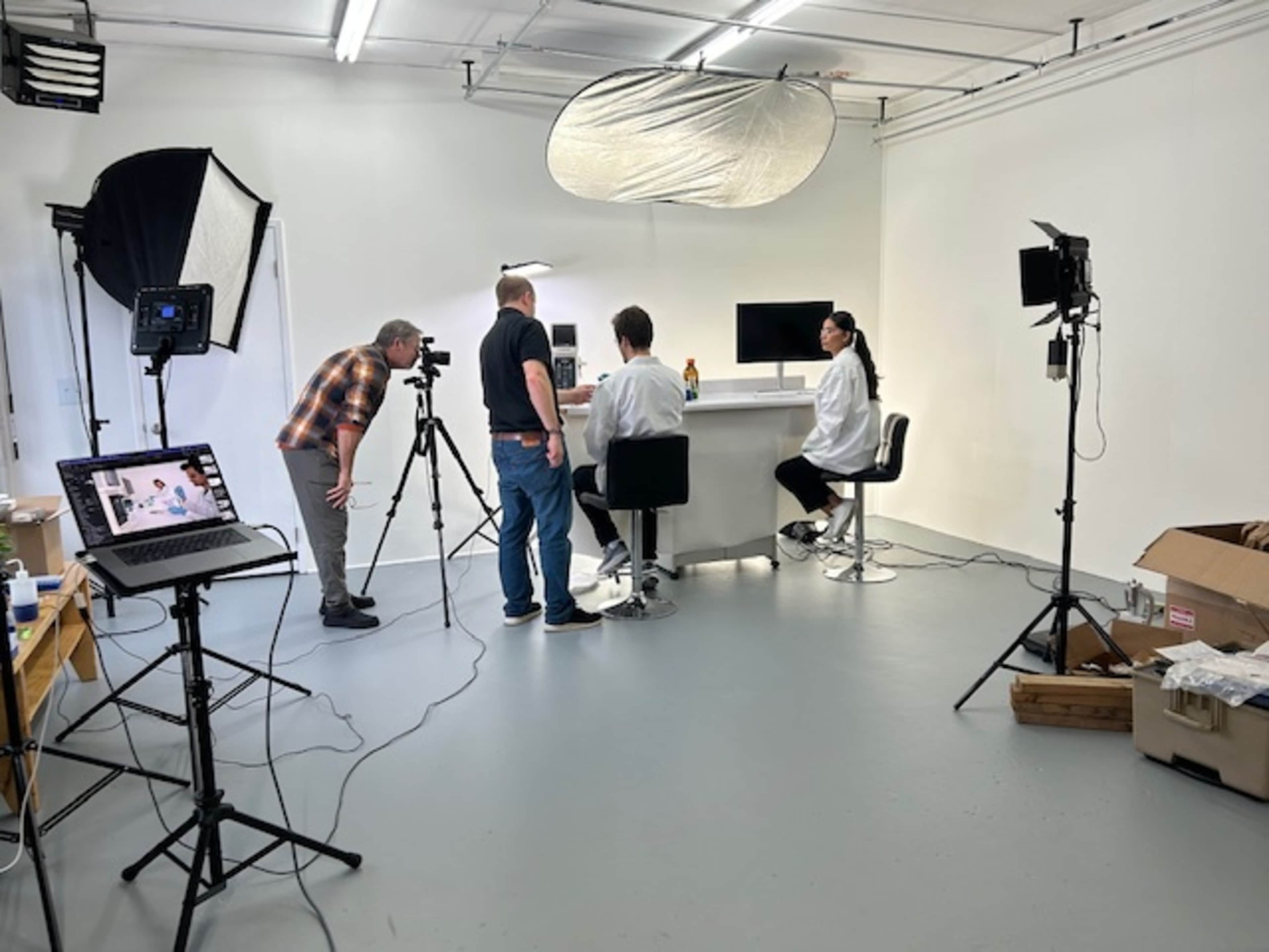 A video production team is set up in a studio, with two individuals seated at a table and cameras positioned to capture the scene.