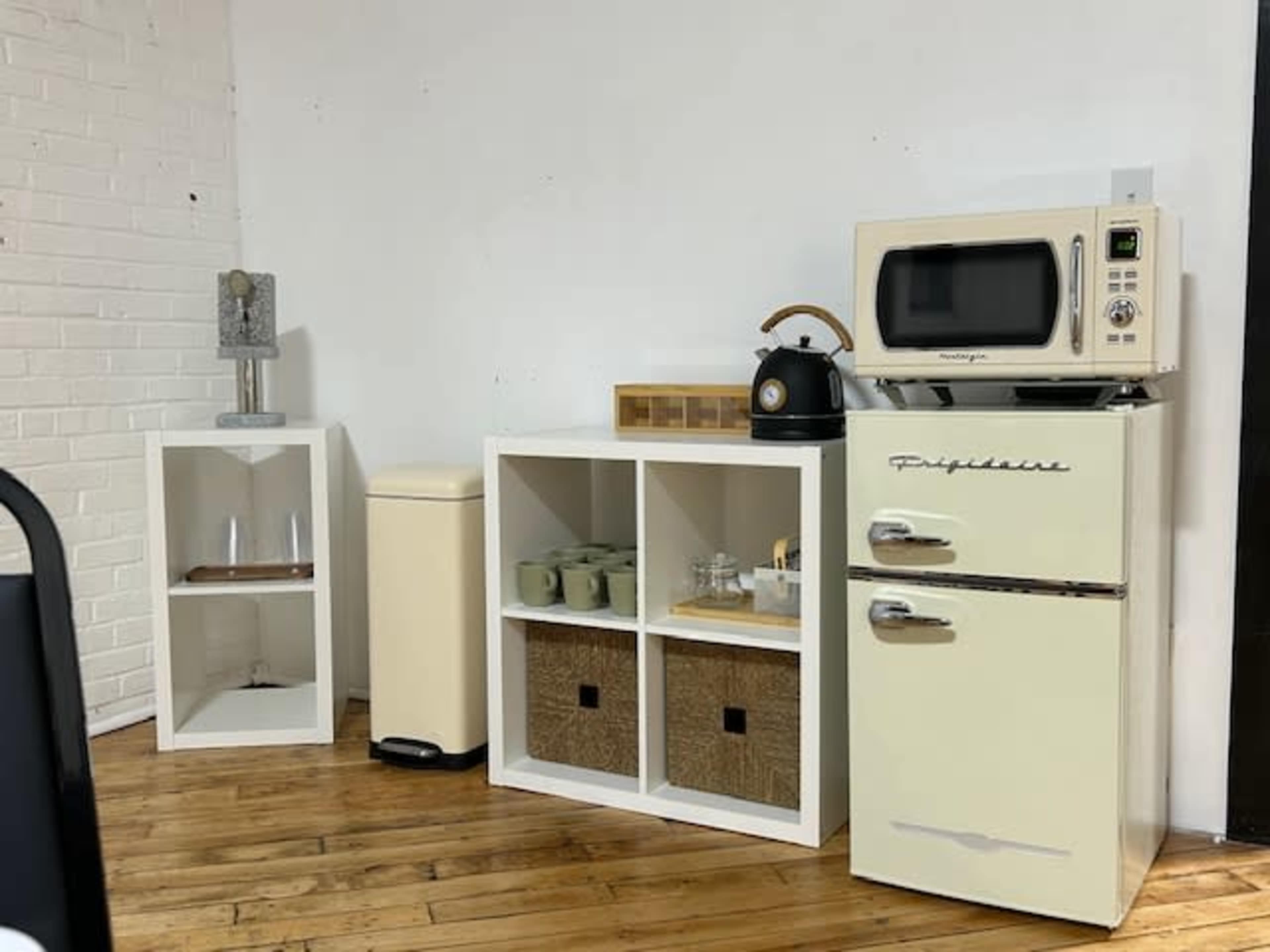 The image features a minimalist kitchen corner with a cream-colored refrigerator, a microwave, a kettle, and storage shelves containing baskets and kitchenware against a white brick wall.