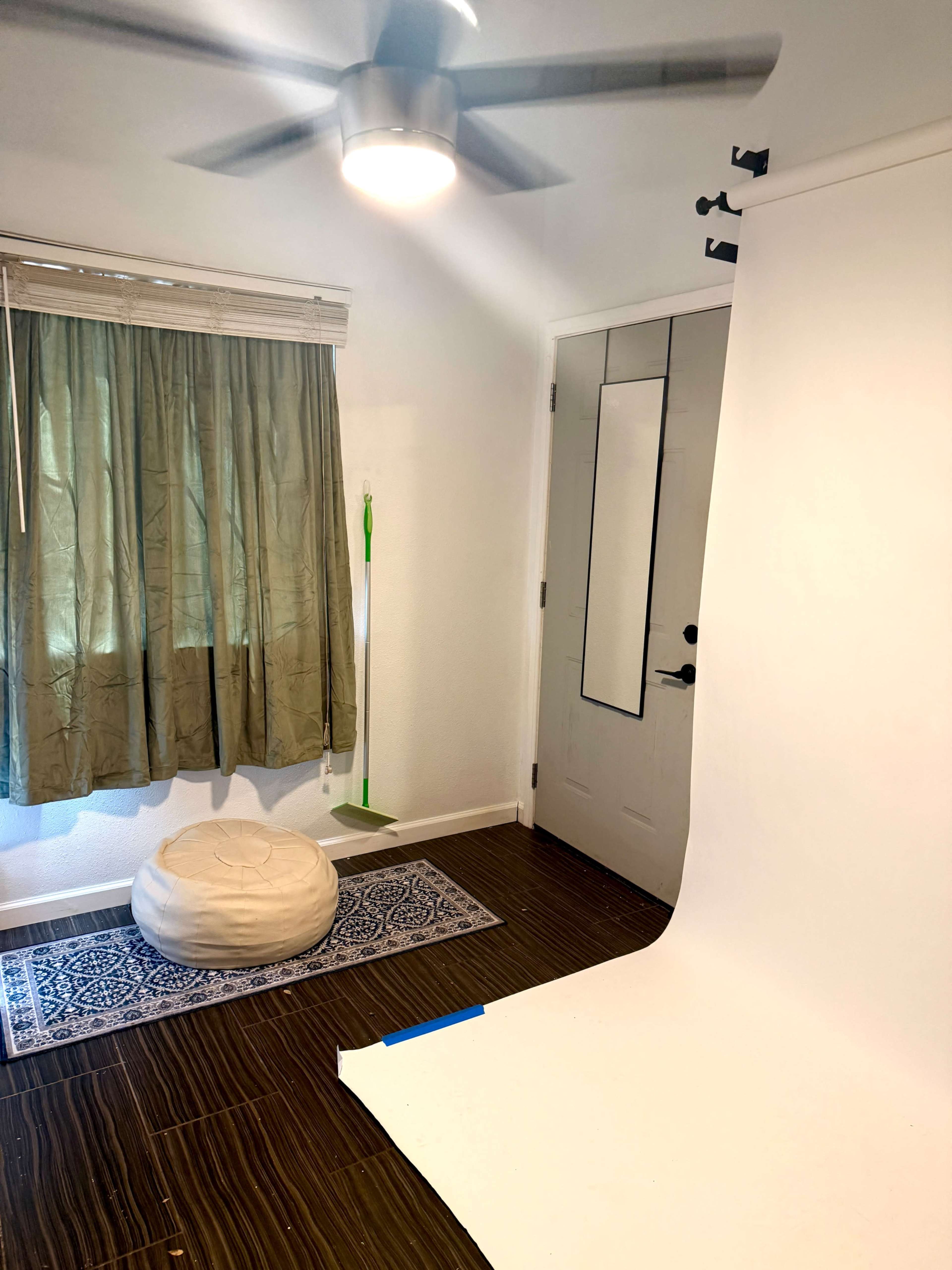 The image shows a room with a white backdrop, a green curtain, a door with a mirror, a broom against the wall, and a white pouf on a patterned rug.