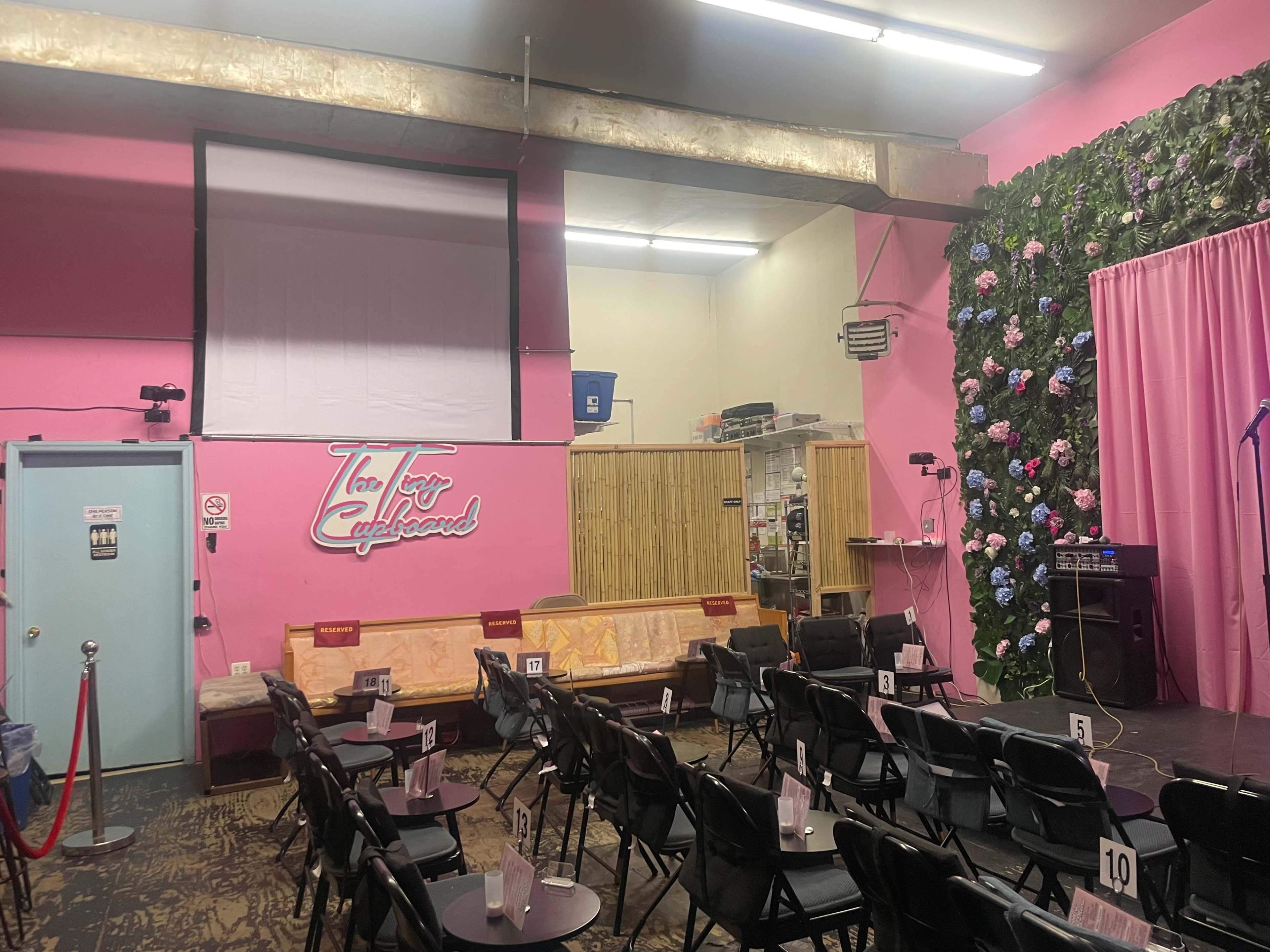 A small performance venue with pink walls, a stage area adorned with artificial flowers, and black chairs arranged for an audience.