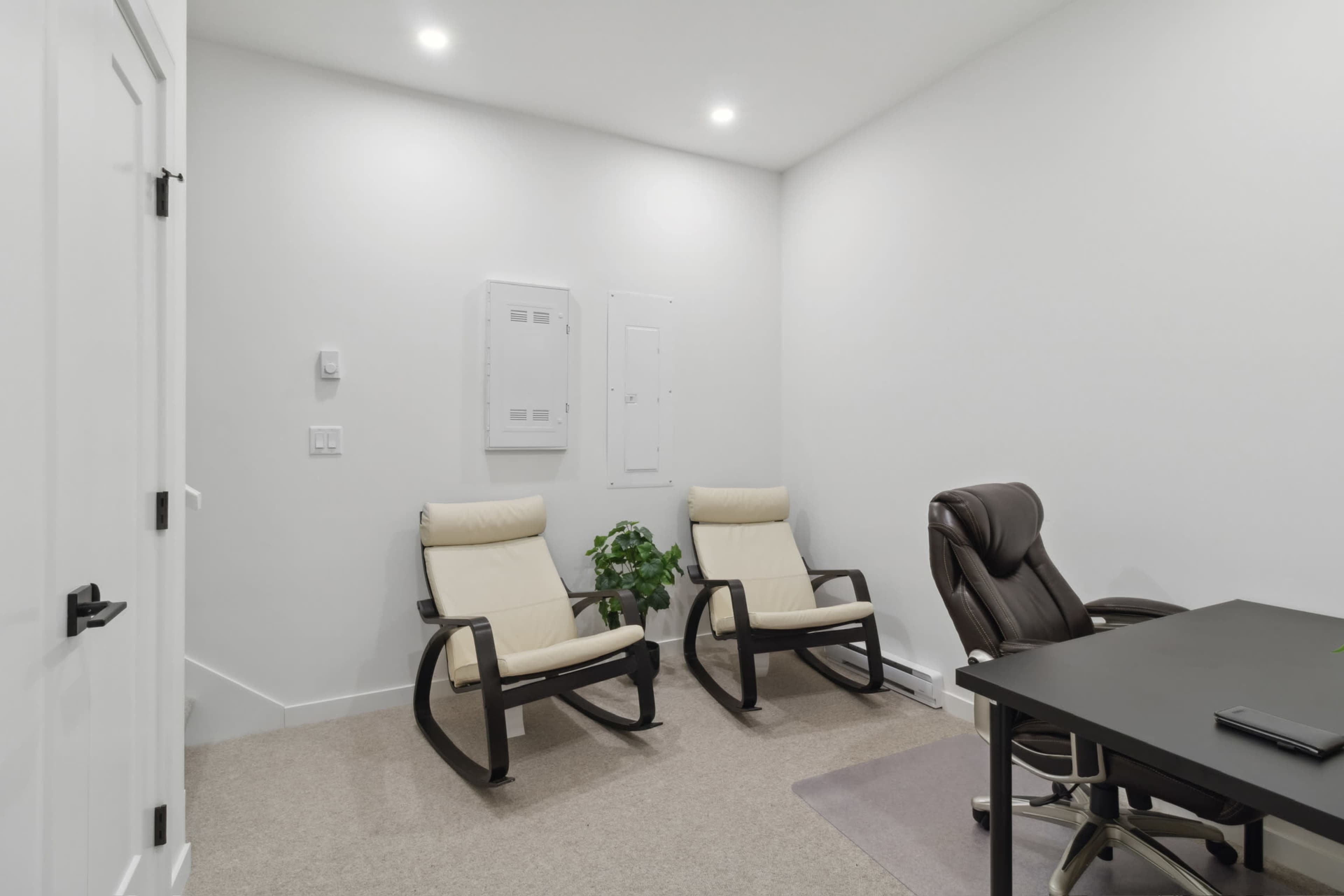The image shows a small, well-lit room with two reclining chairs, a potted plant, and a black desk with a swivel chair.