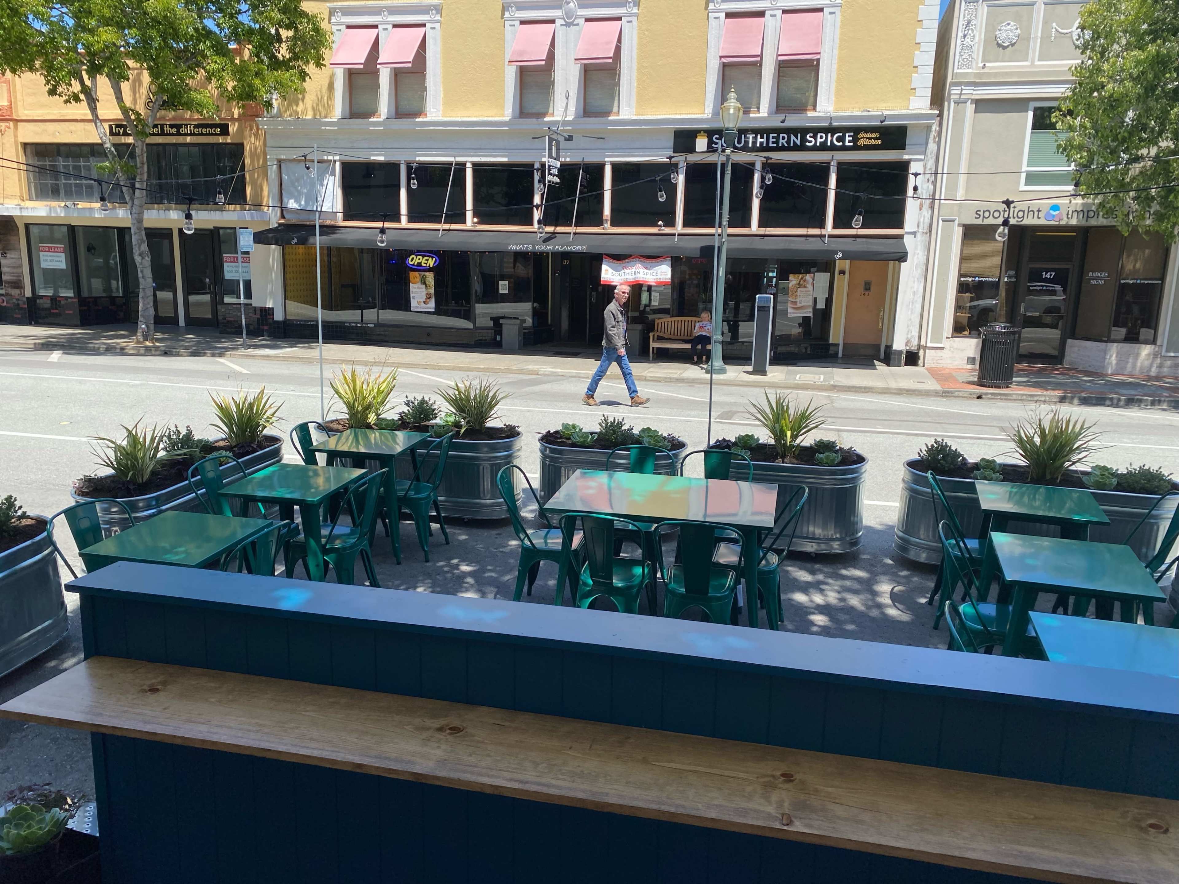 A row of green tables and chairs are set outside a restaurant on a street lined with buildings.
