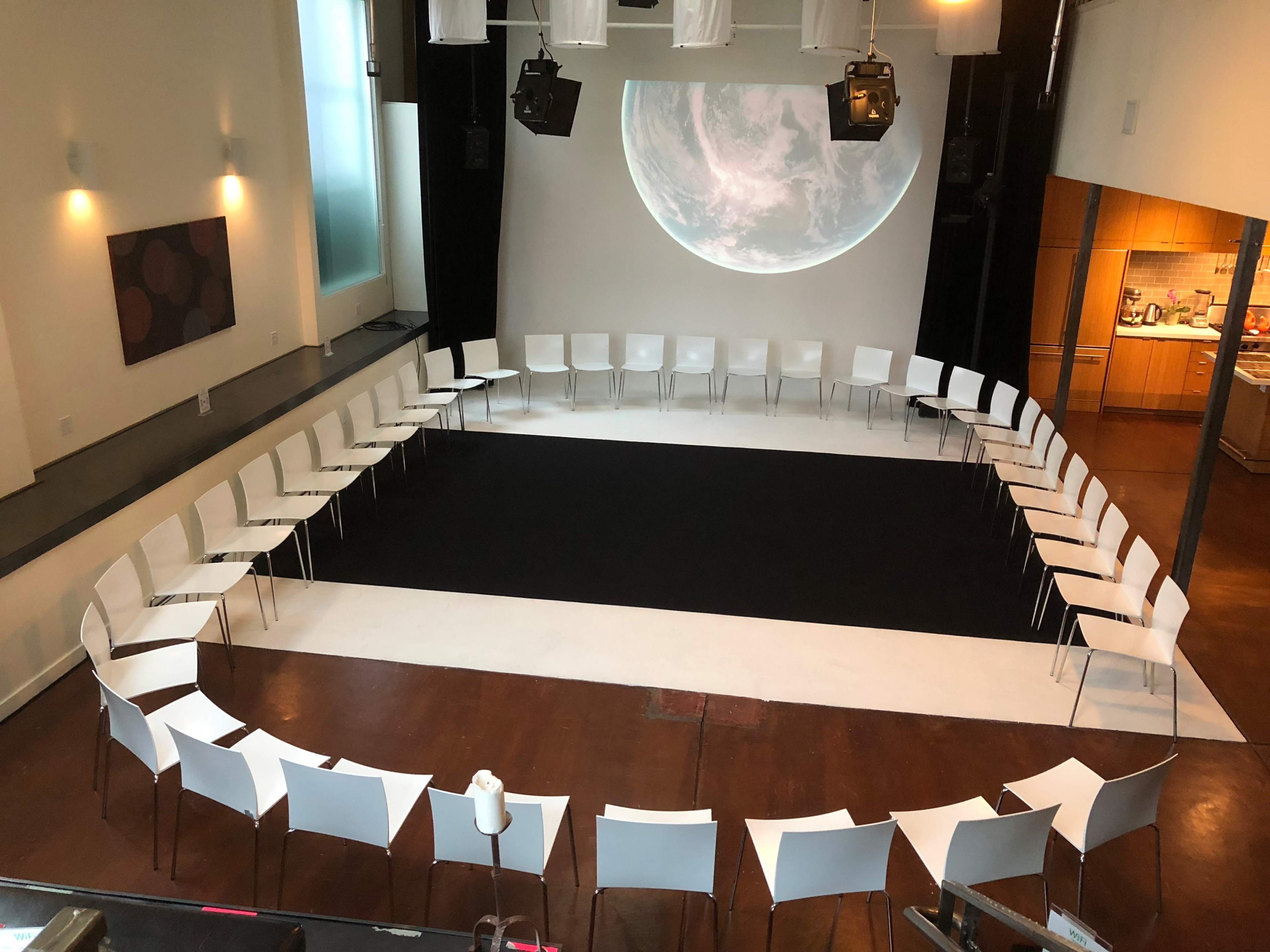 A spacious room arranged with a circular set of white chairs facing a blank screen displaying a large circular graphic, with a kitchen area visible in the background.