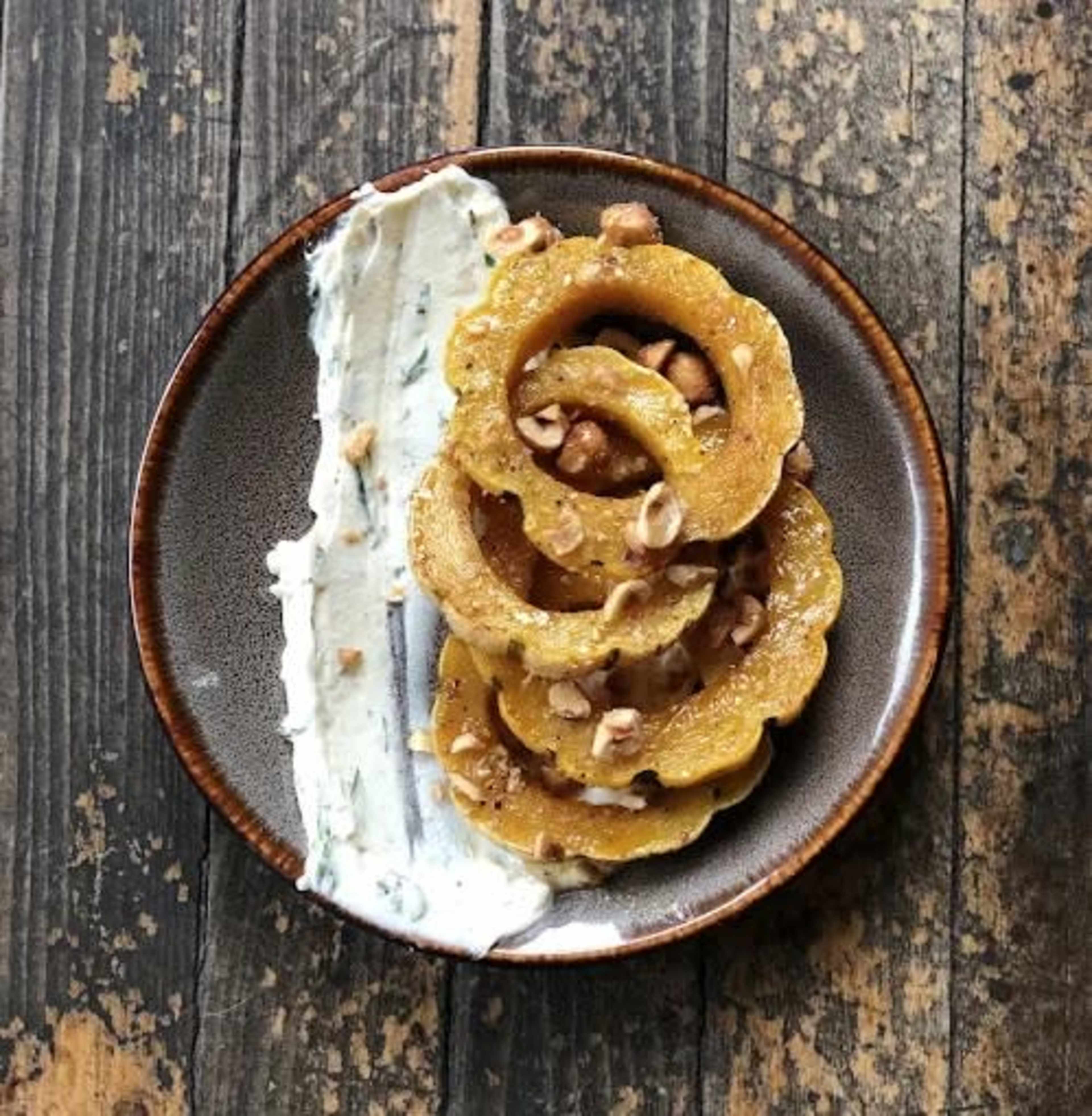 A plate holds circular slices of squash topped with chopped nuts beside a dollop of a creamy sauce.