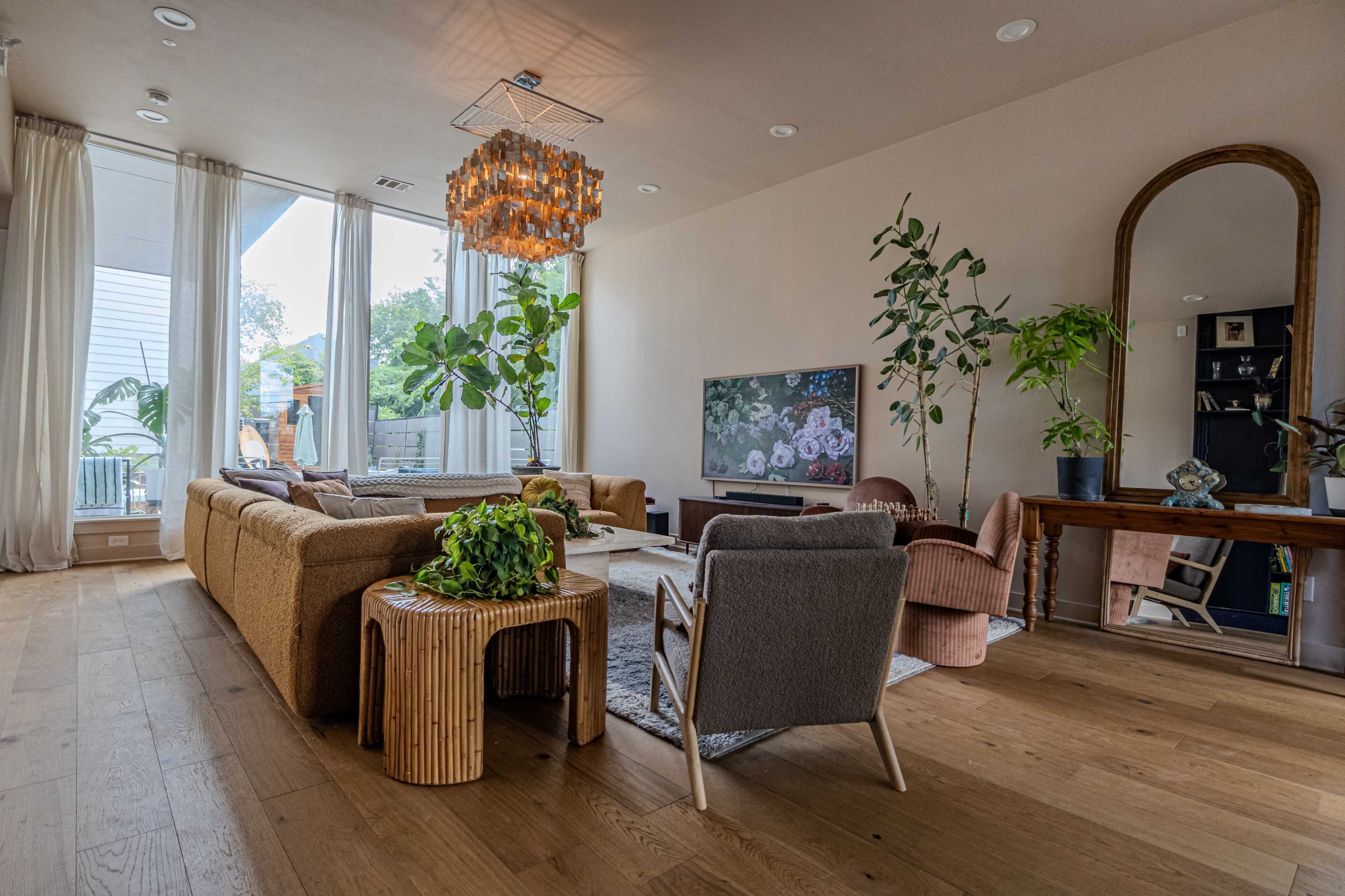 The image shows a modern living room featuring a mix of seating options, plants, a large wall-mounted TV, and a decorative chandelier.