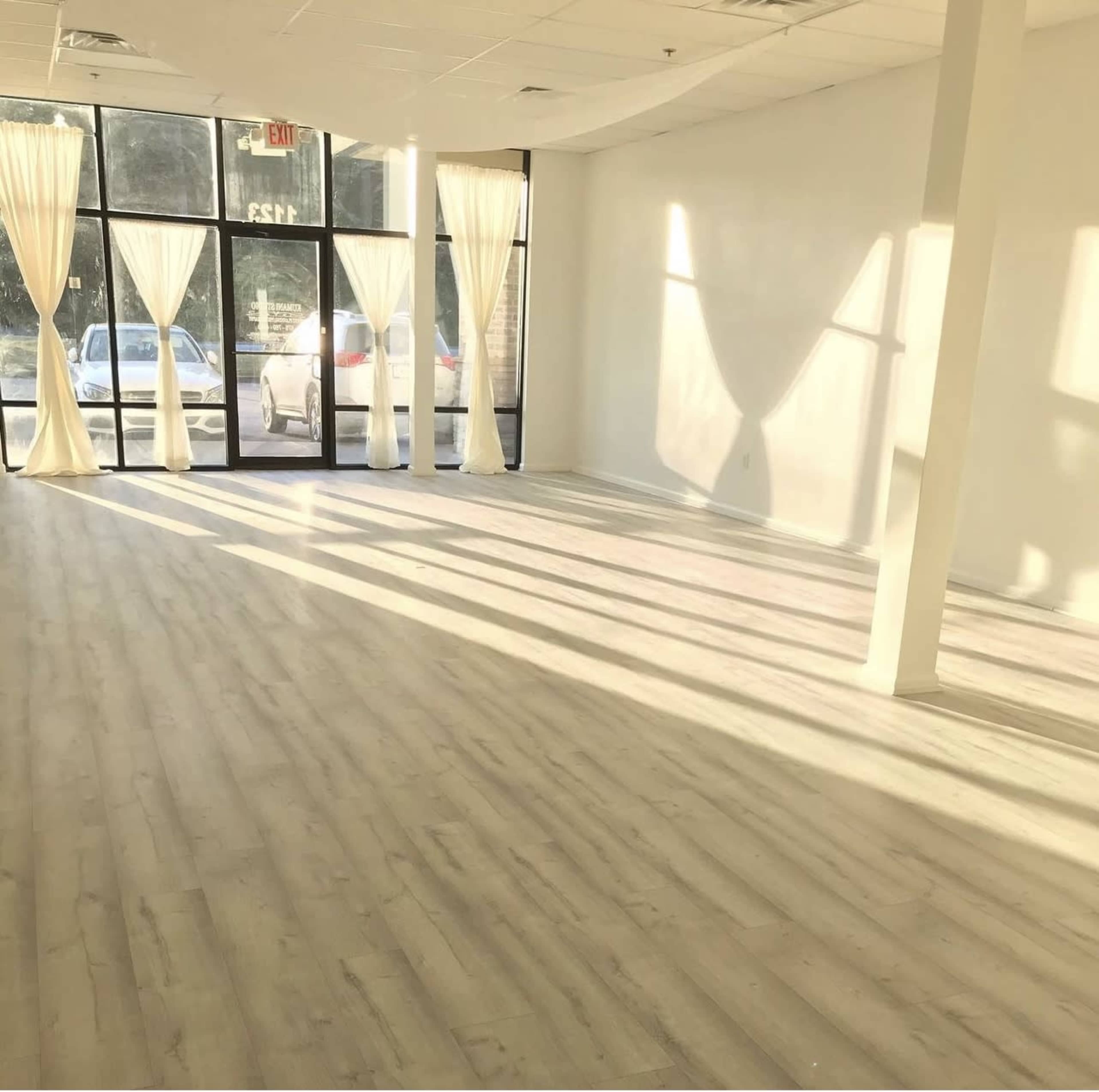 A bright, empty room with large windows draped with white curtains, casting long shadows on a light-colored wooden floor.