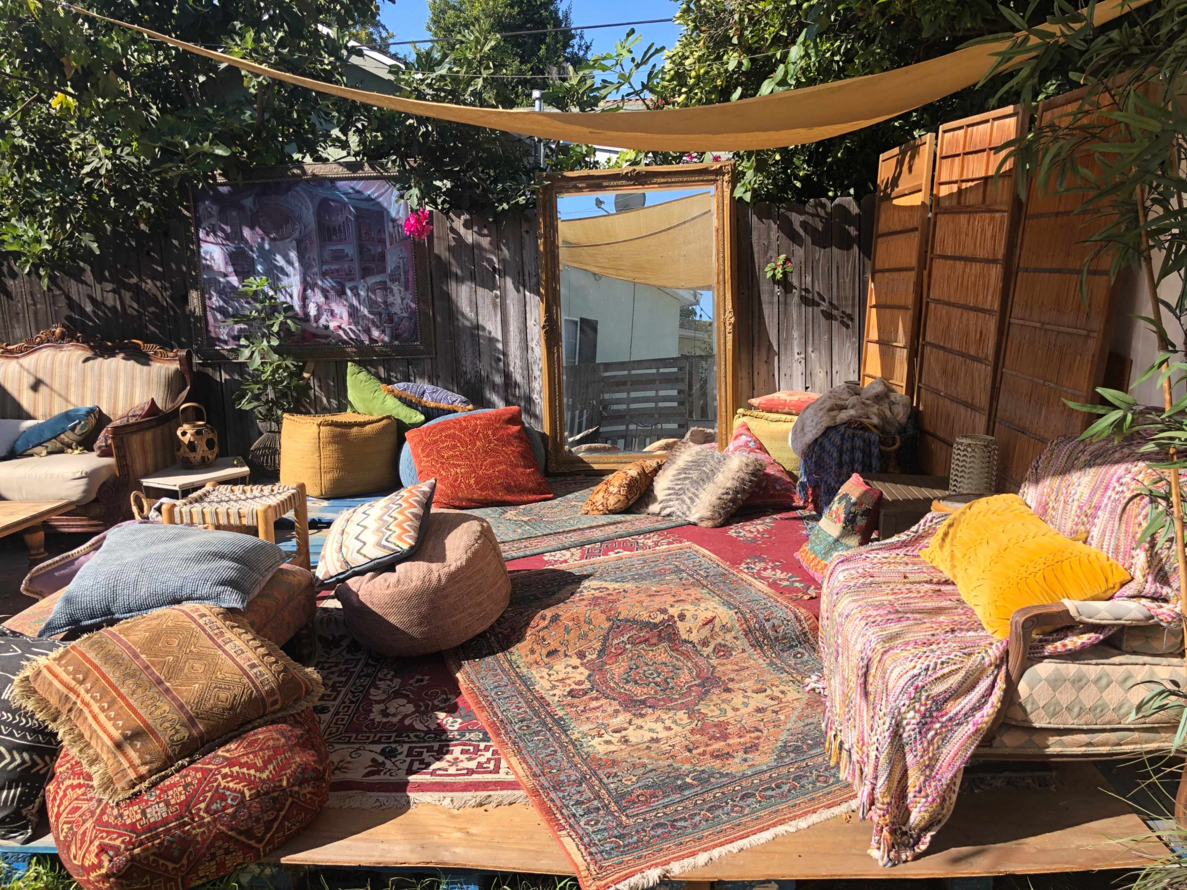 The scene features a cozy outdoor seating area adorned with various cushions, rugs, and a large mirror surrounded by greenery.