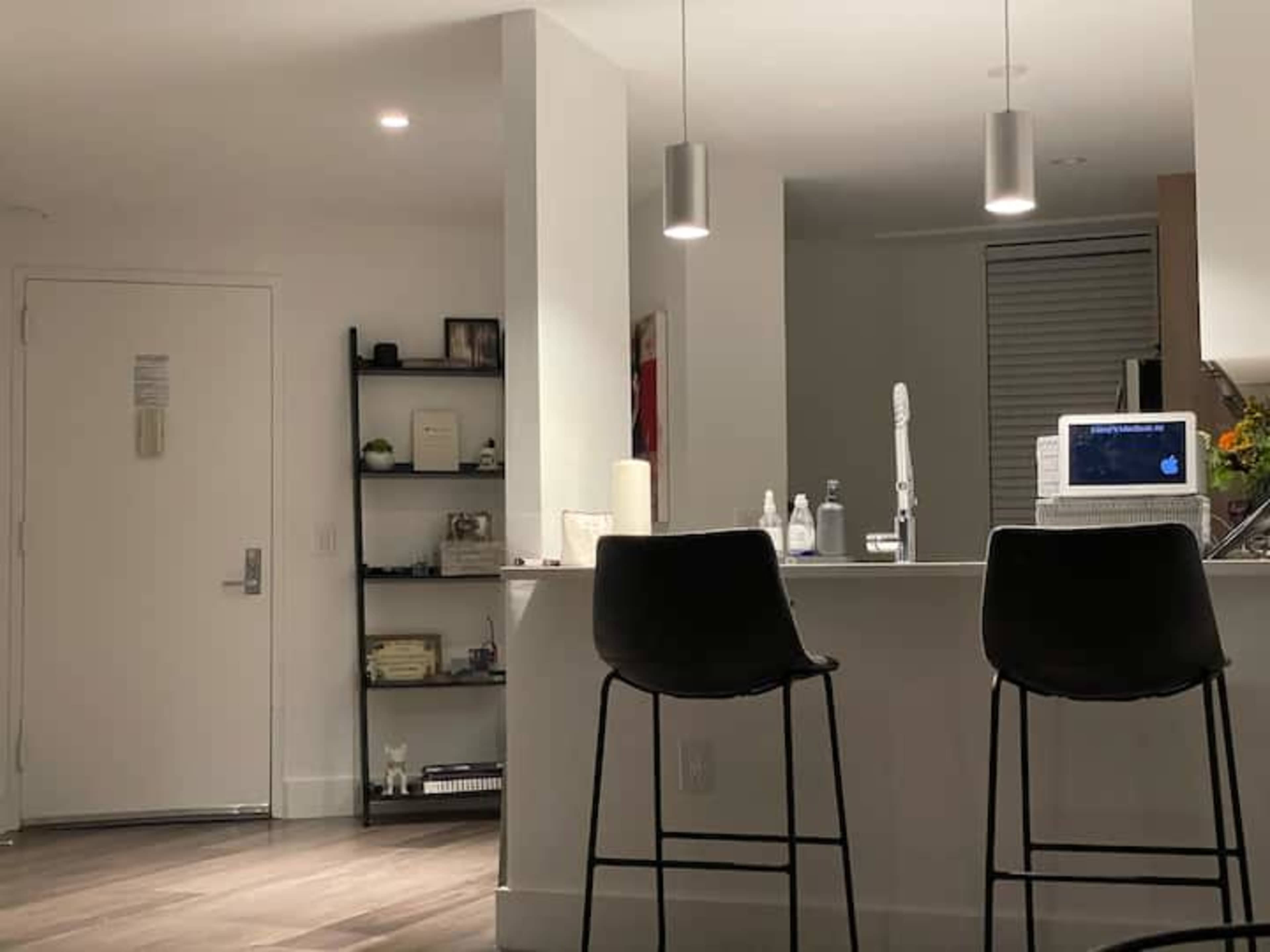 A modern kitchen with two black bar stools at a countertop, a computer on the counter, and a door visible to the left.