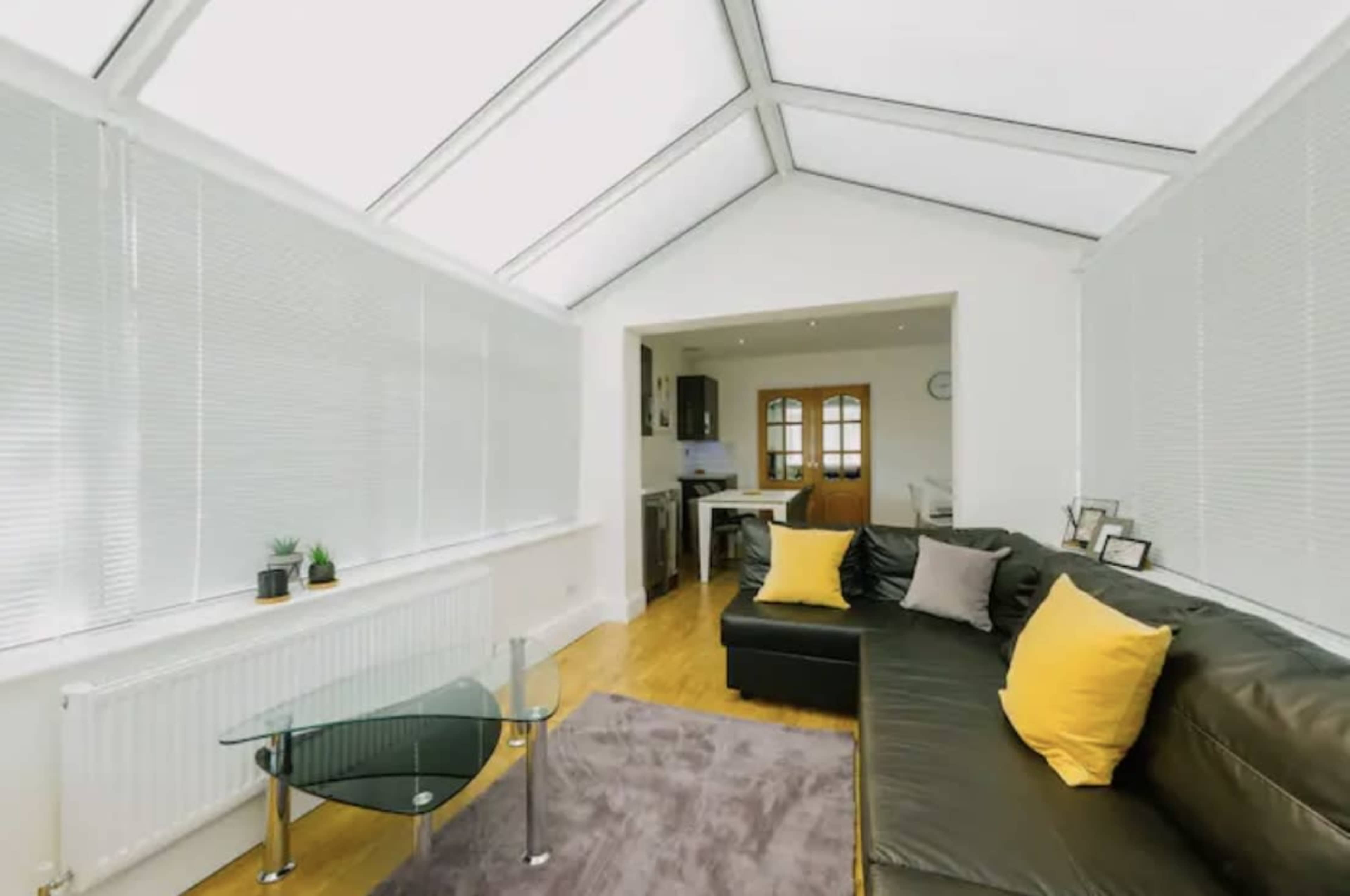 A sunroom features a black leather sofa with yellow cushions, a glass coffee table, and large windows with white blinds overlooking a kitchen area.