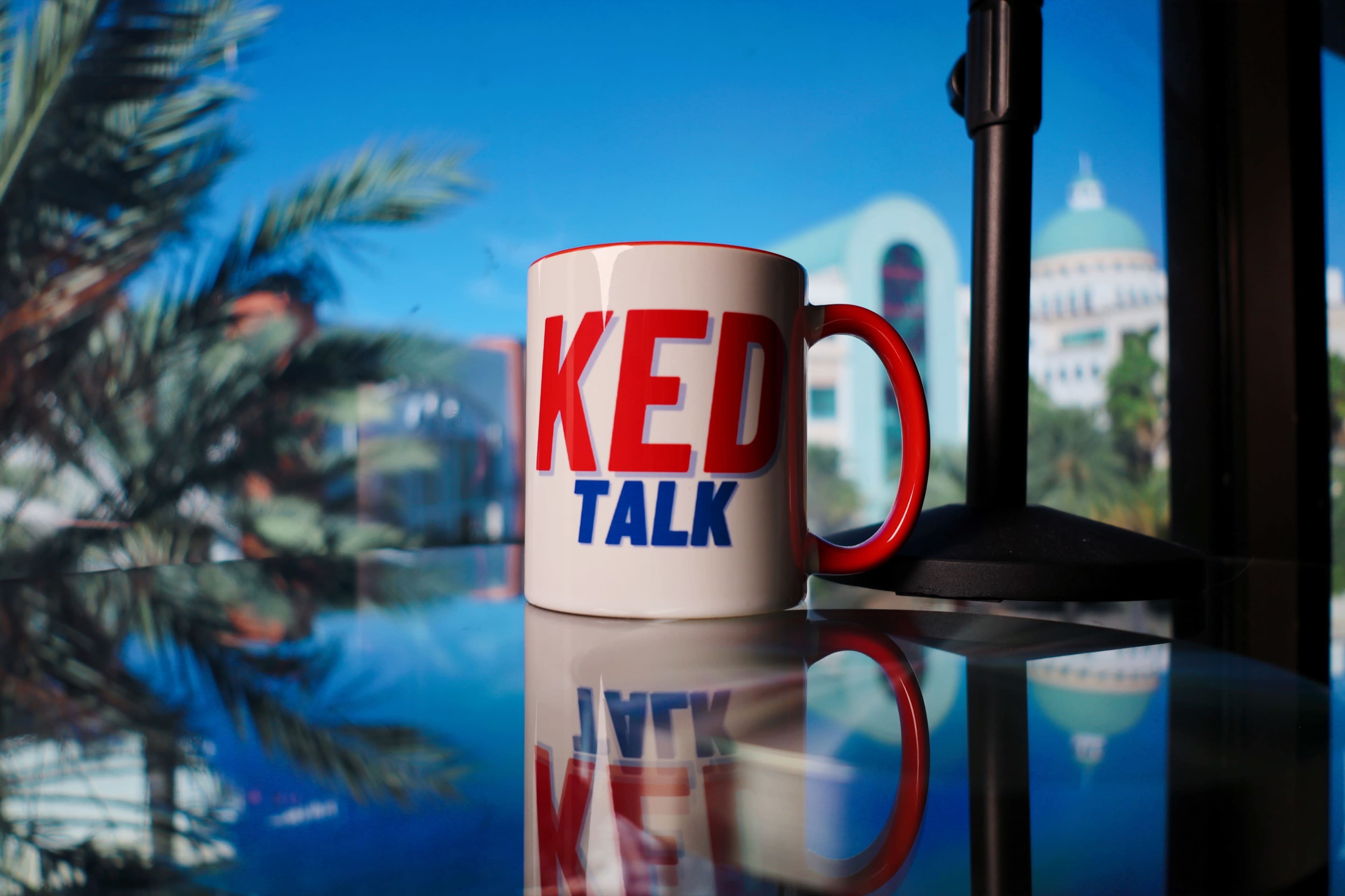A coffee mug featuring the words "KED TALK" sits on a reflective surface with palm trees and a building visible in the background.