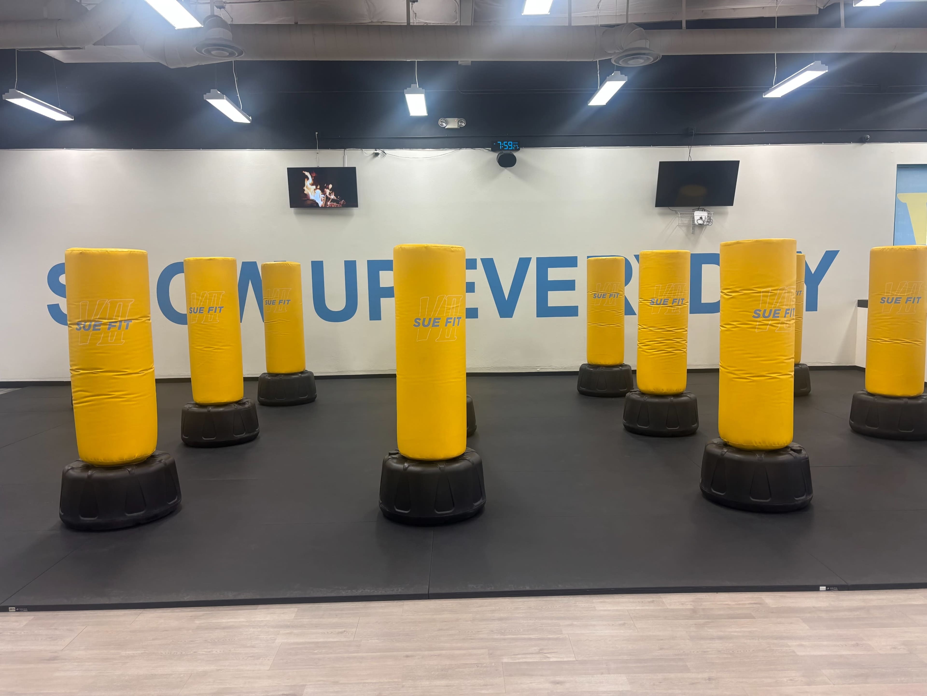 The image shows a fitness studio with multiple yellow punching bags arranged on a black mat floor against a blue wall with the words "SWIM EVERY DAY."