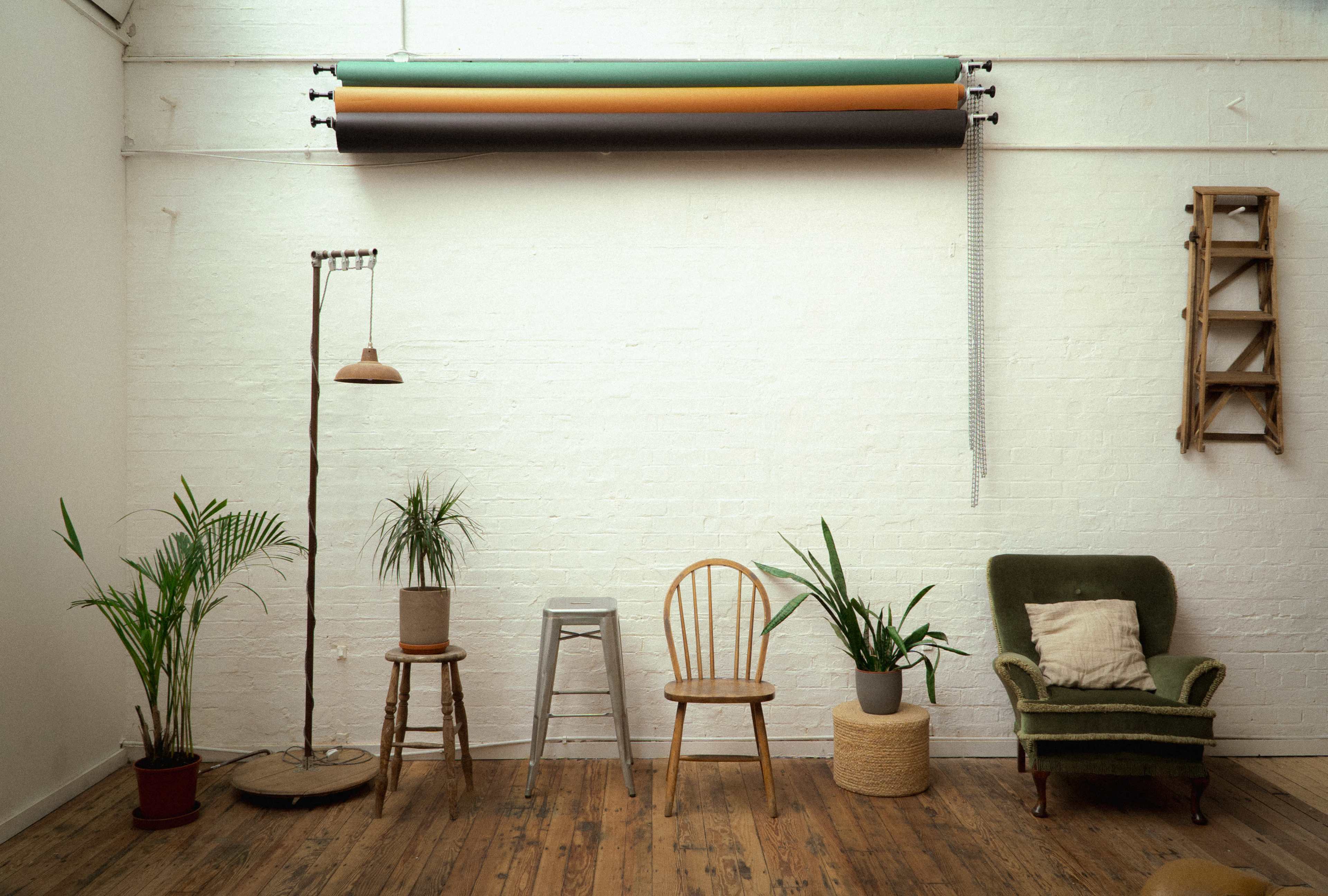 A minimalistic interior space with a variety of potted plants, a green armchair, a wooden ladder, and several stools along a white brick wall.