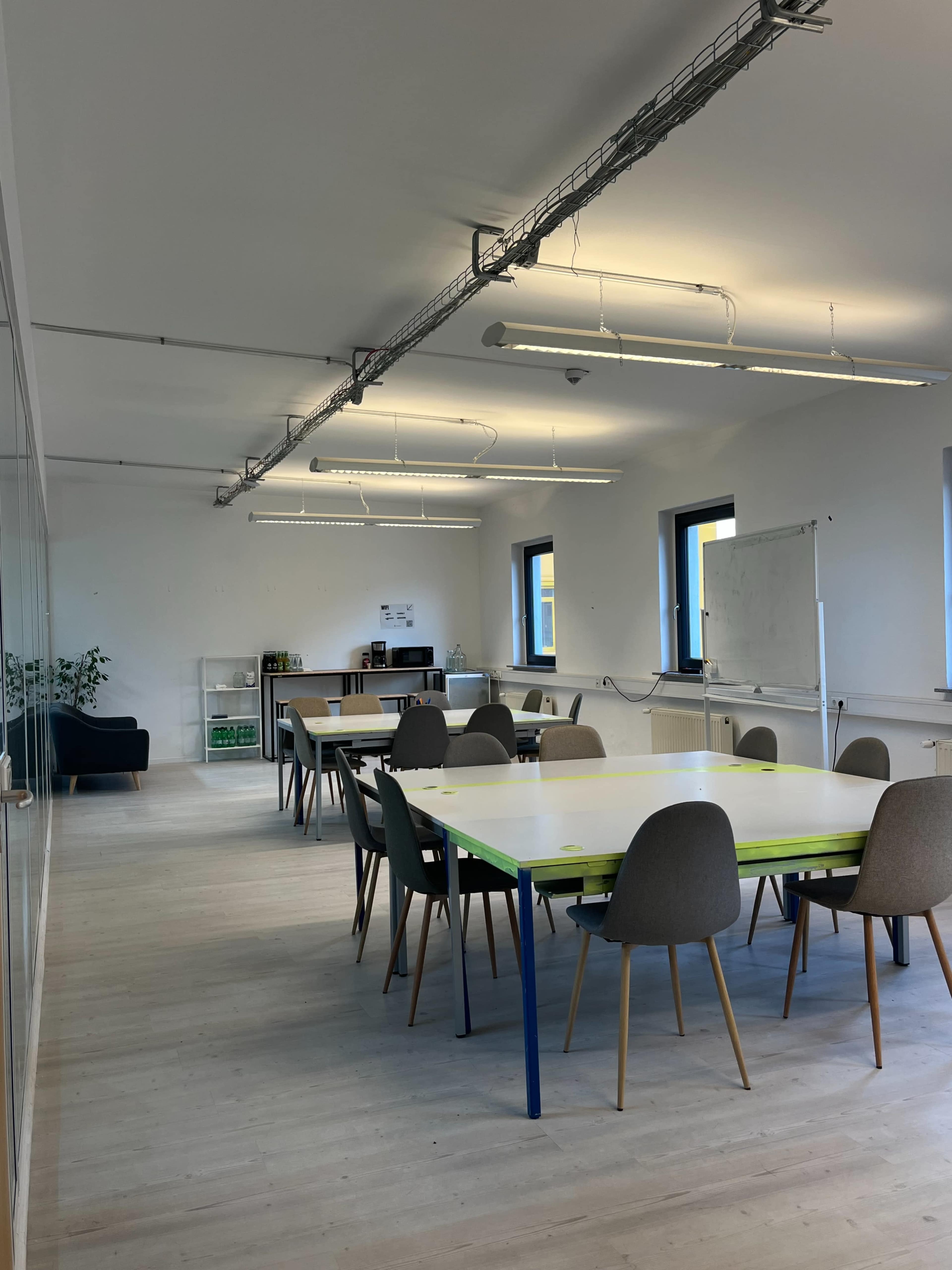 The image shows a modern, spacious meeting room with a long table surrounded by chairs, large windows letting in natural light, and a kitchenette in the background.