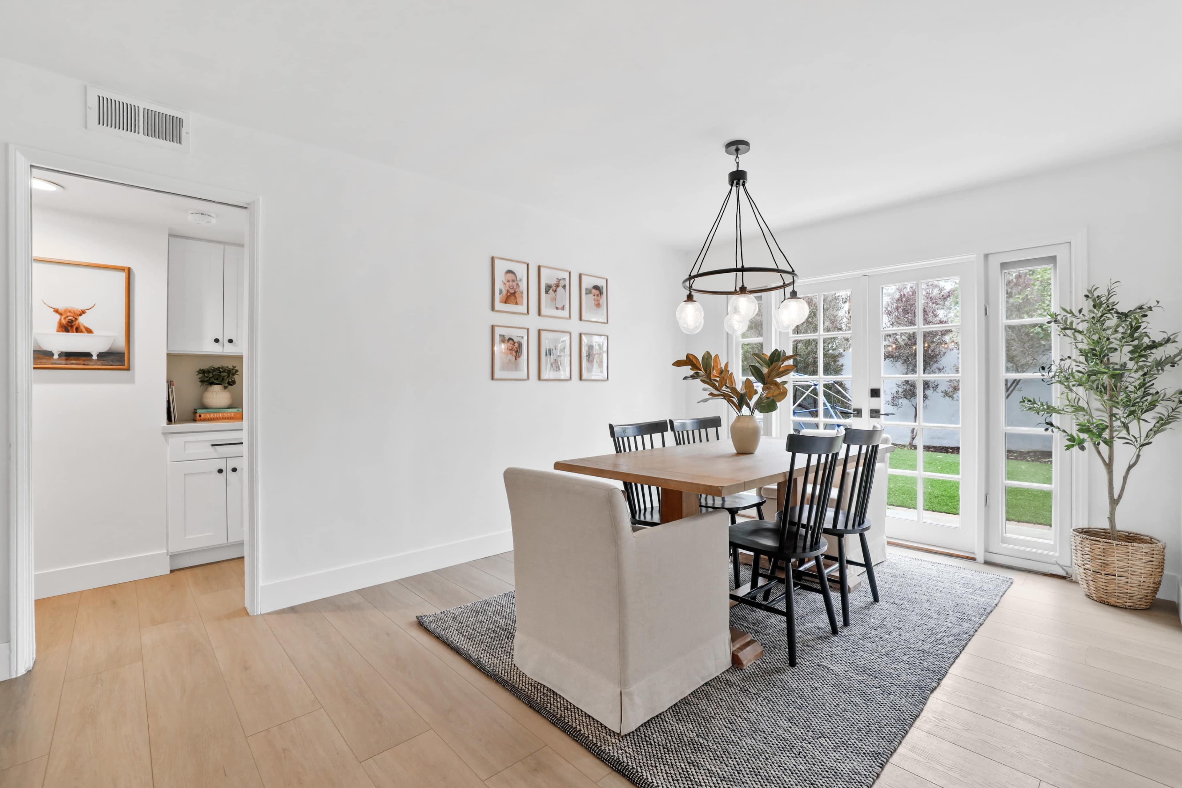 A dining area features a wooden table surrounded by black chairs, with a rug underneath and large windows leading to a garden.
