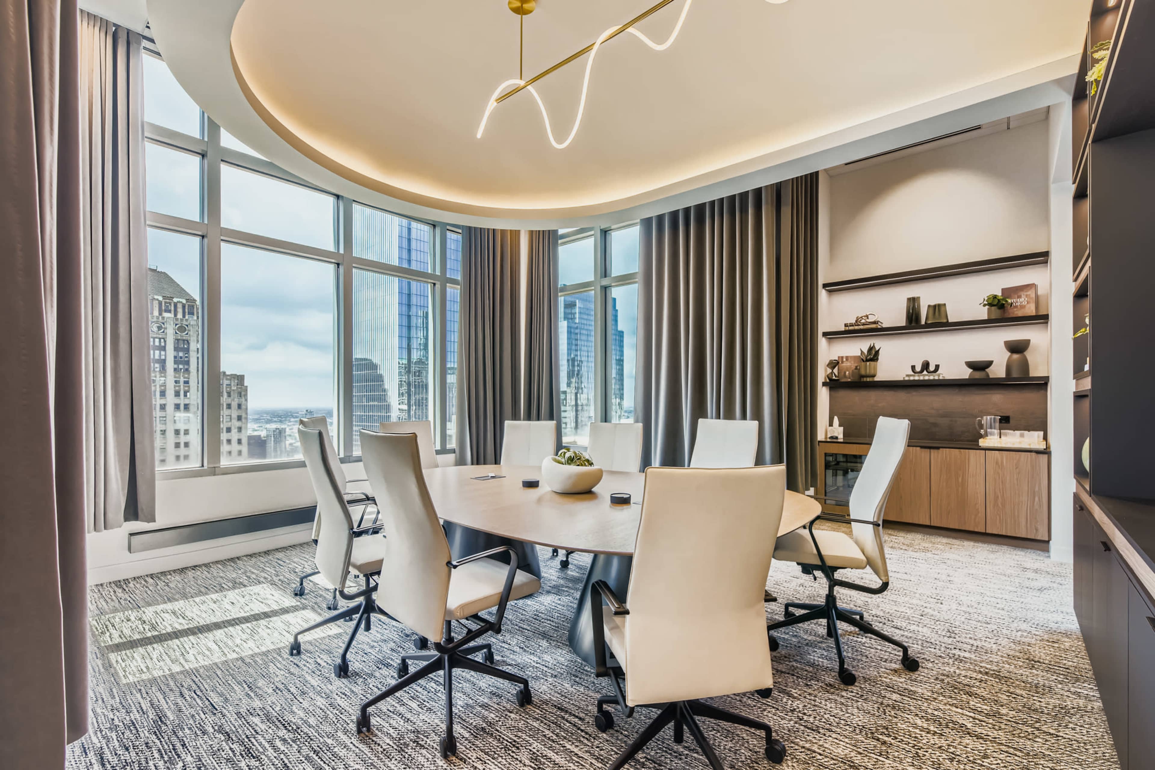 A modern boardroom features a circular table surrounded by six light-colored chairs and large windows overlooking a city skyline.