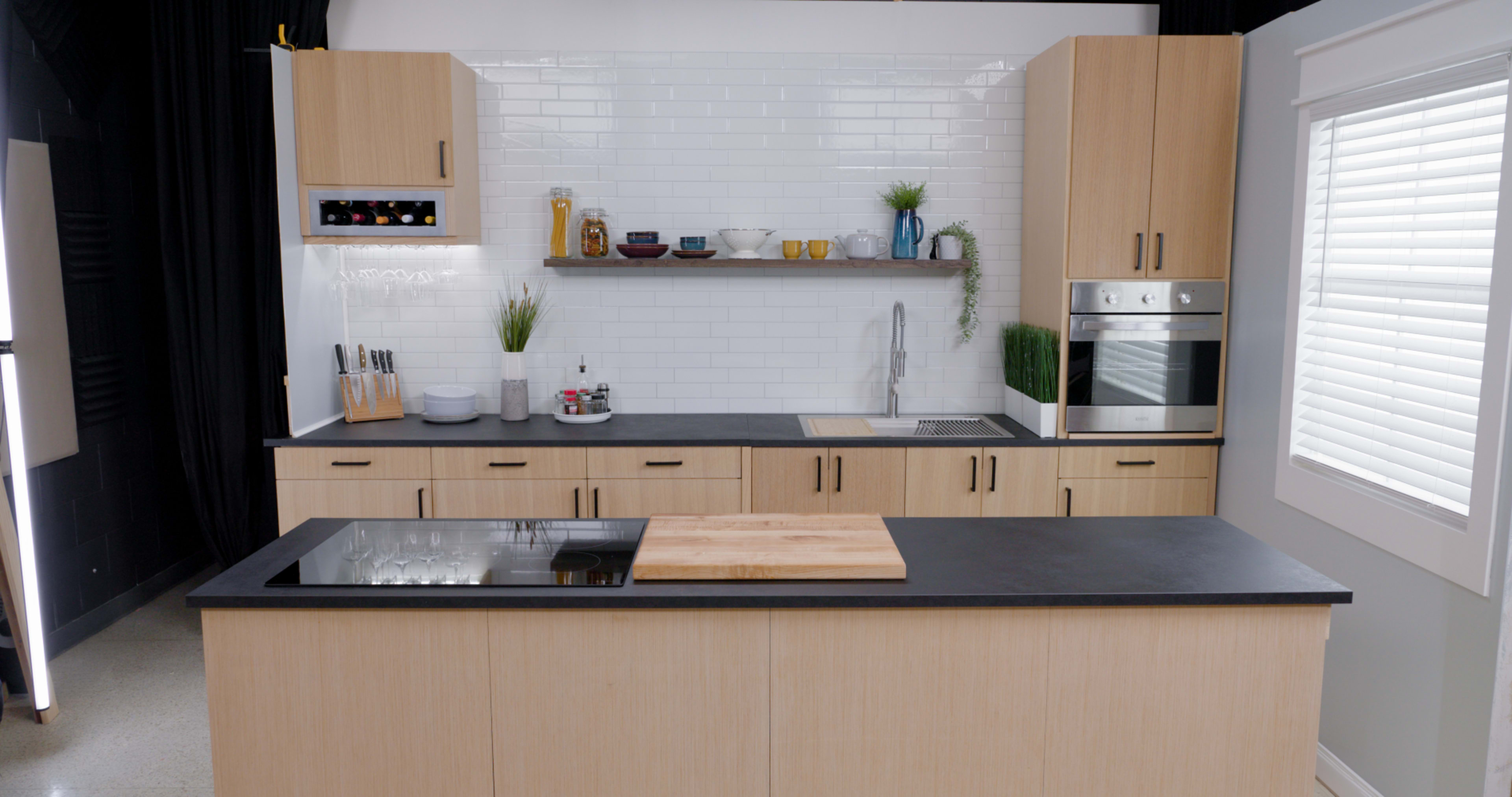 The image shows a modern kitchen with light wood cabinetry, a black countertop, an island with a wooden cutting board, and a minimalist design featuring various kitchen items on shelves.