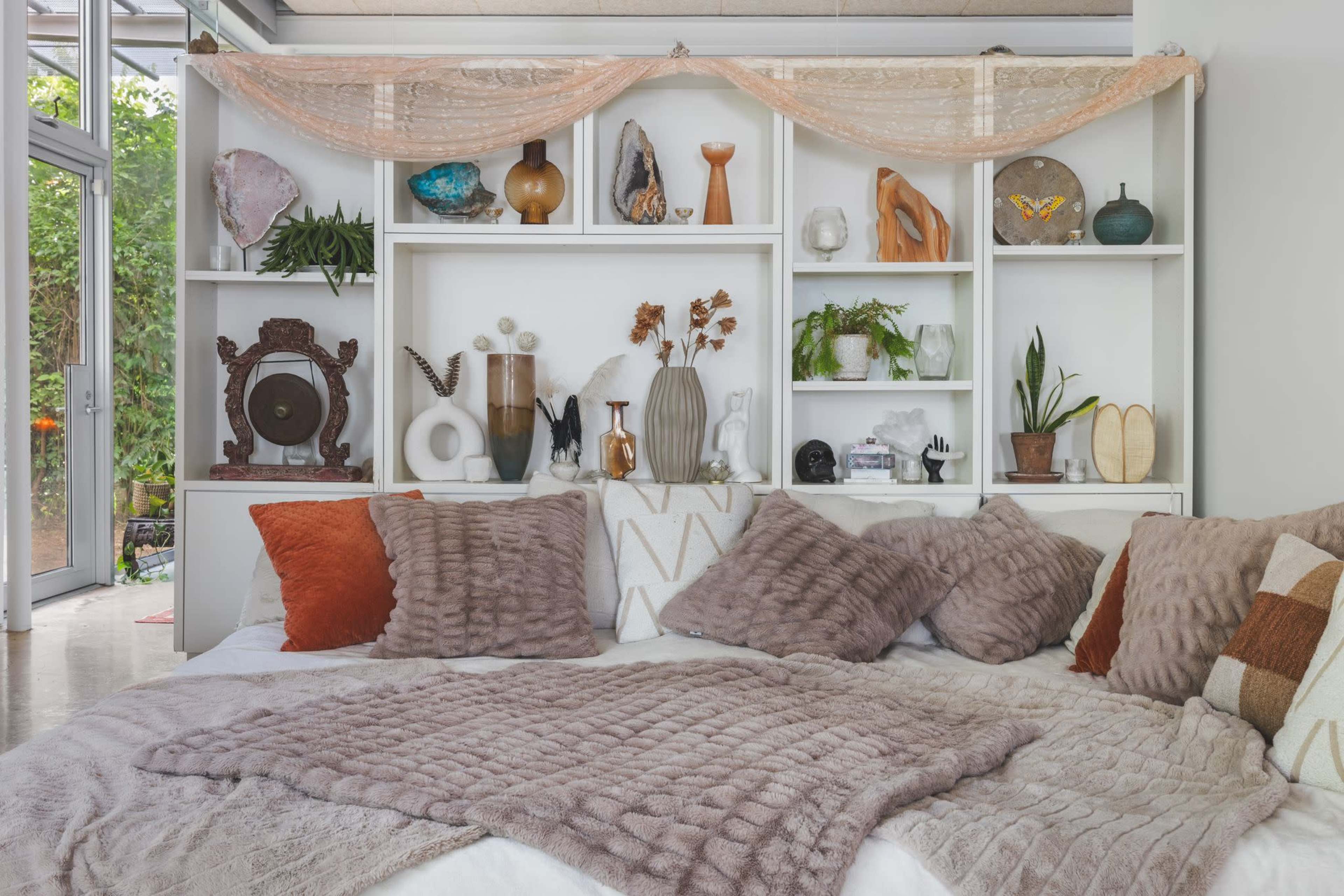 A cozy seating area features a bed with various textured pillows, draped with a soft blanket, in front of a decorative shelf filled with plants and art objects.