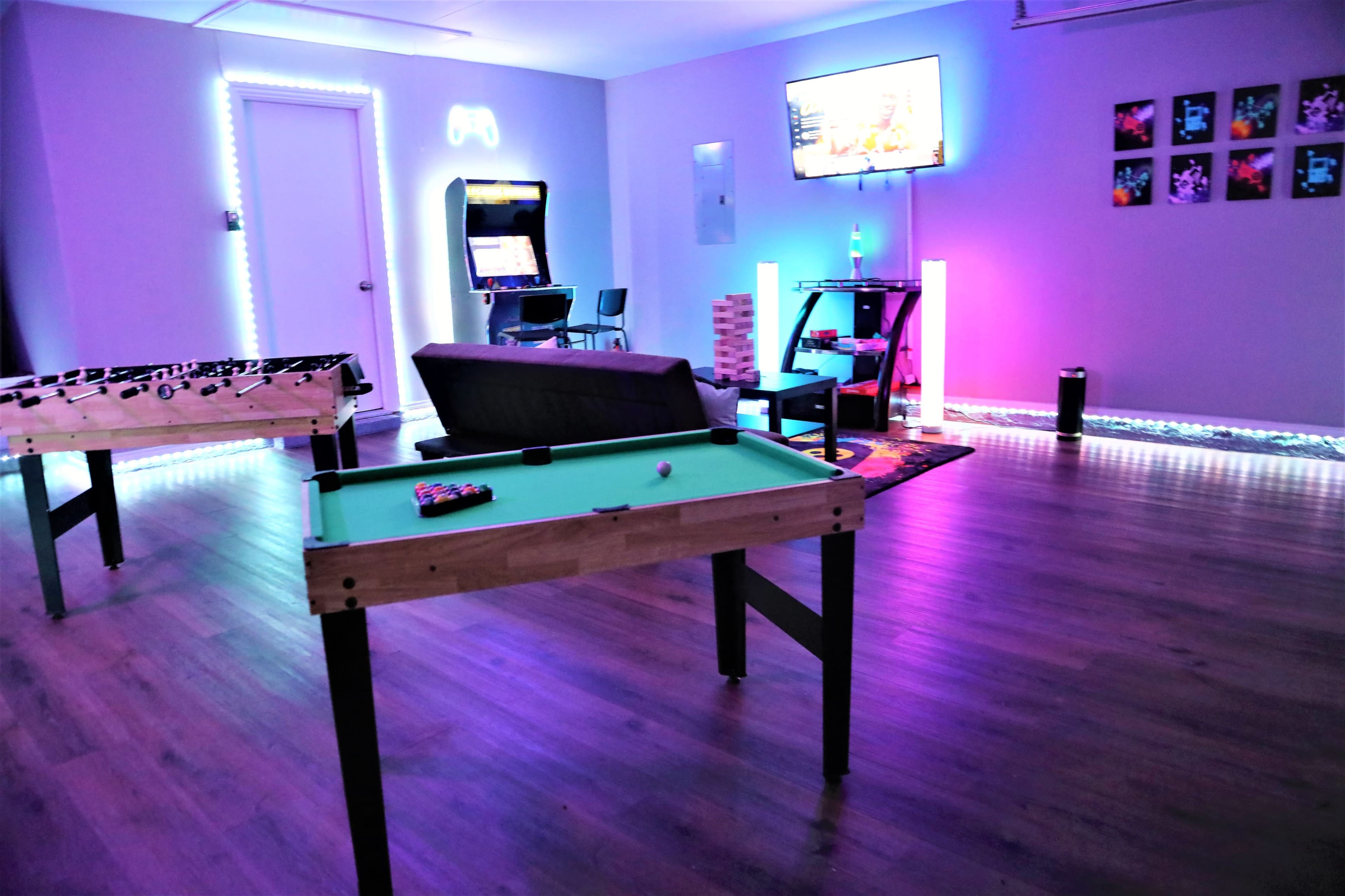 A game room features a pool table, foosball table, arcade machine, and a TV, all illuminated with colorful lighting.