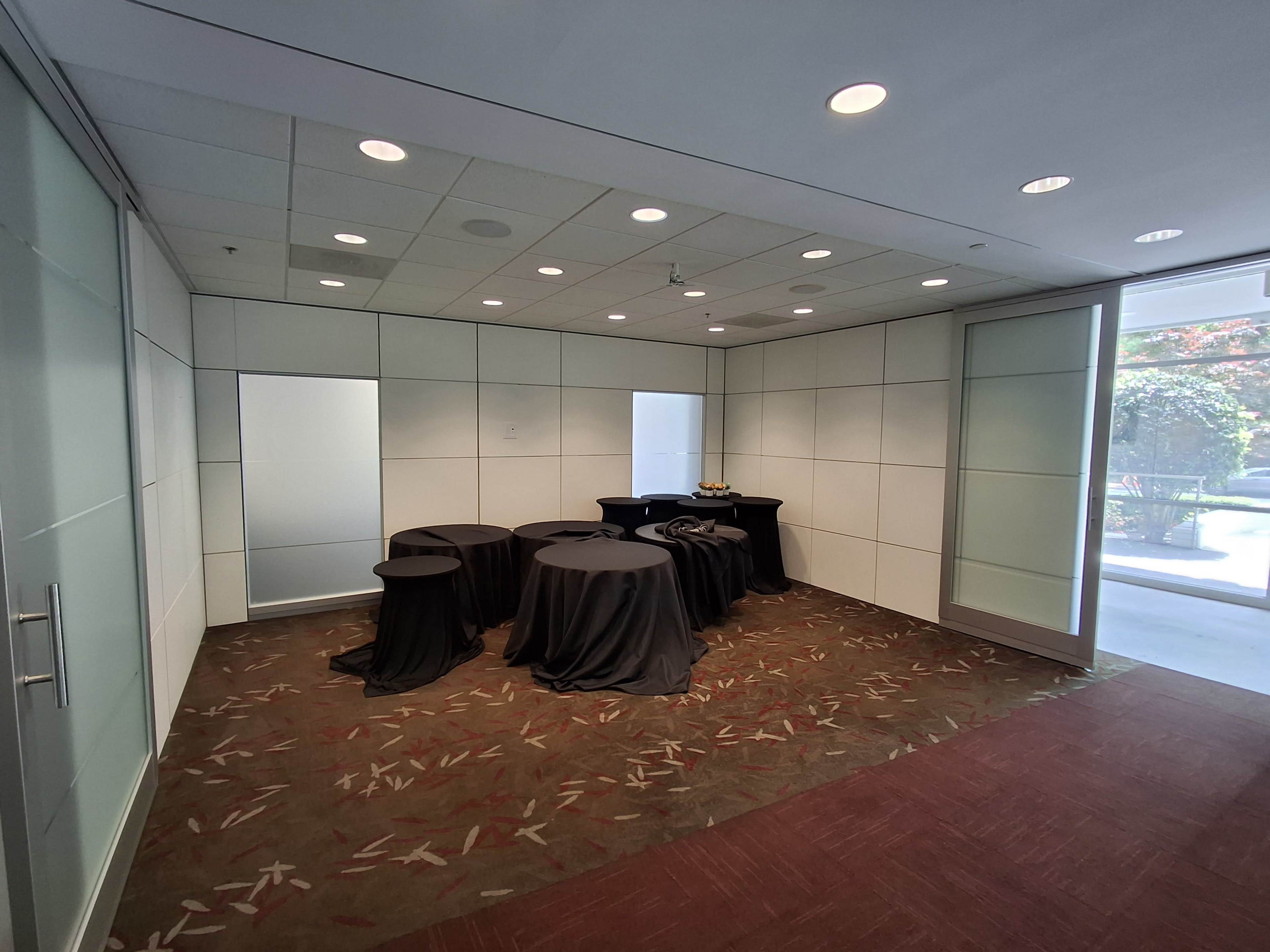 The image shows a modern conference room with several small tables covered in black cloth, surrounded by light-colored walls and large windows.