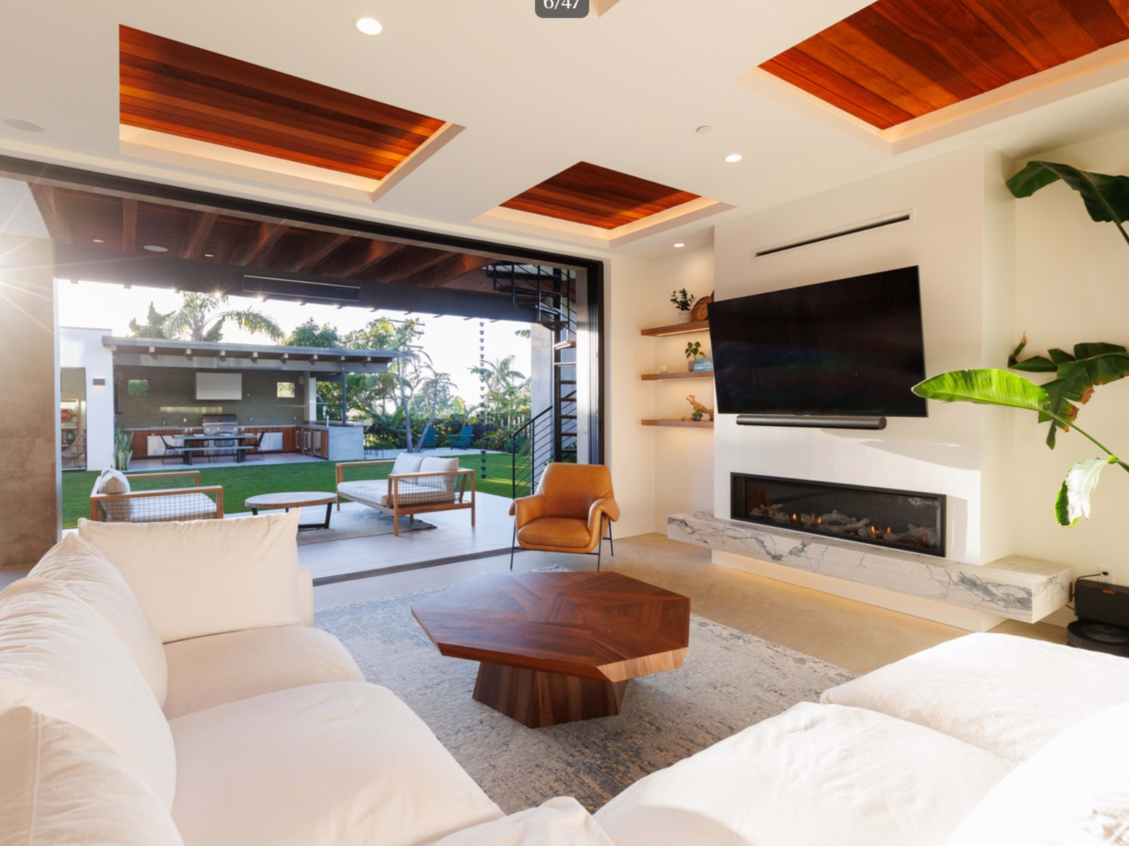 The modern living room features a large sectional sofa, a wooden coffee table, and a flat-screen TV mounted on the wall, with wide doors opening to a backyard patio.