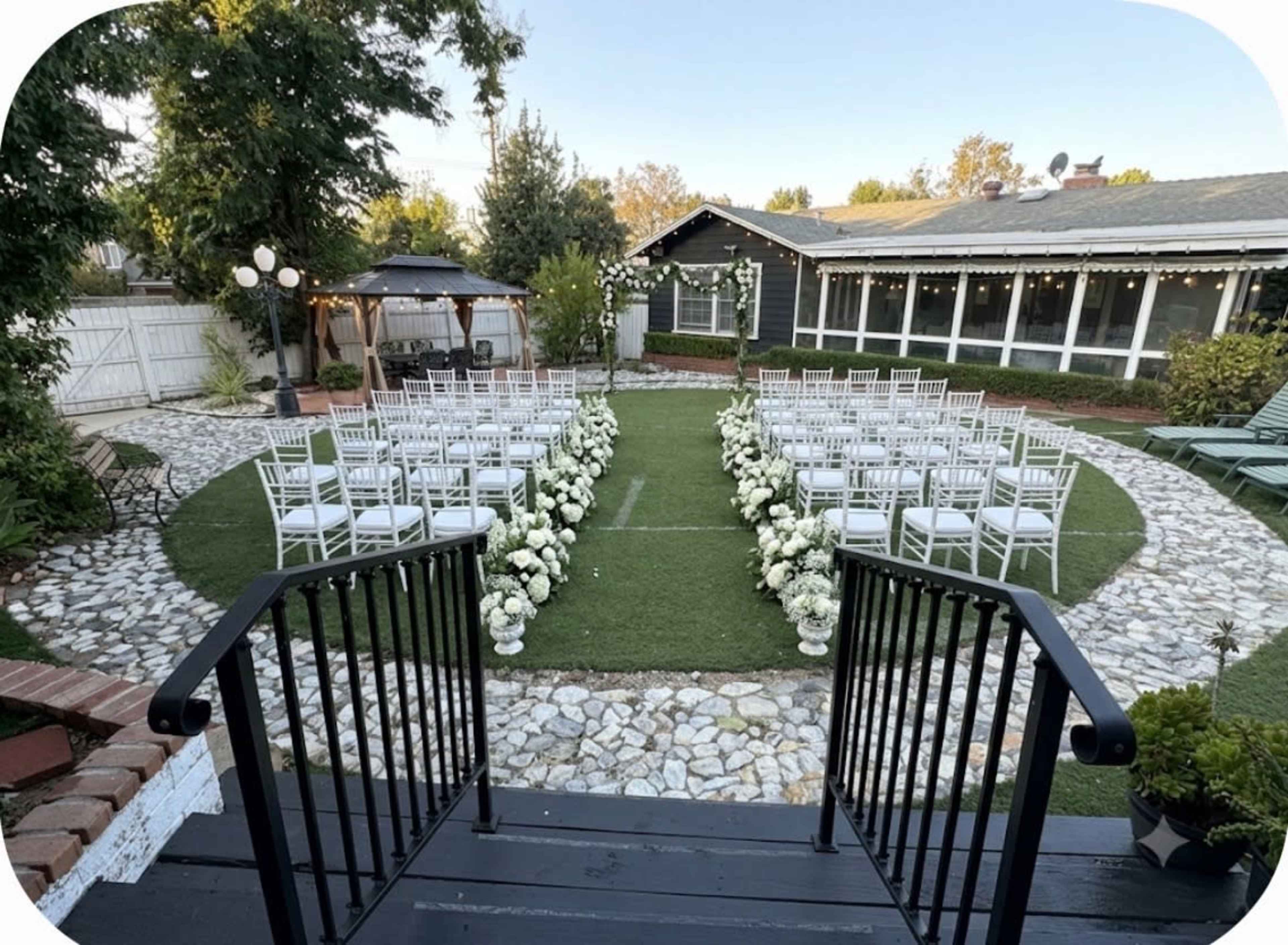 An outdoor wedding ceremony setup features neatly arranged white chairs on a grassy area surrounded by flower arrangements in white vases, with a gazebo in the background.