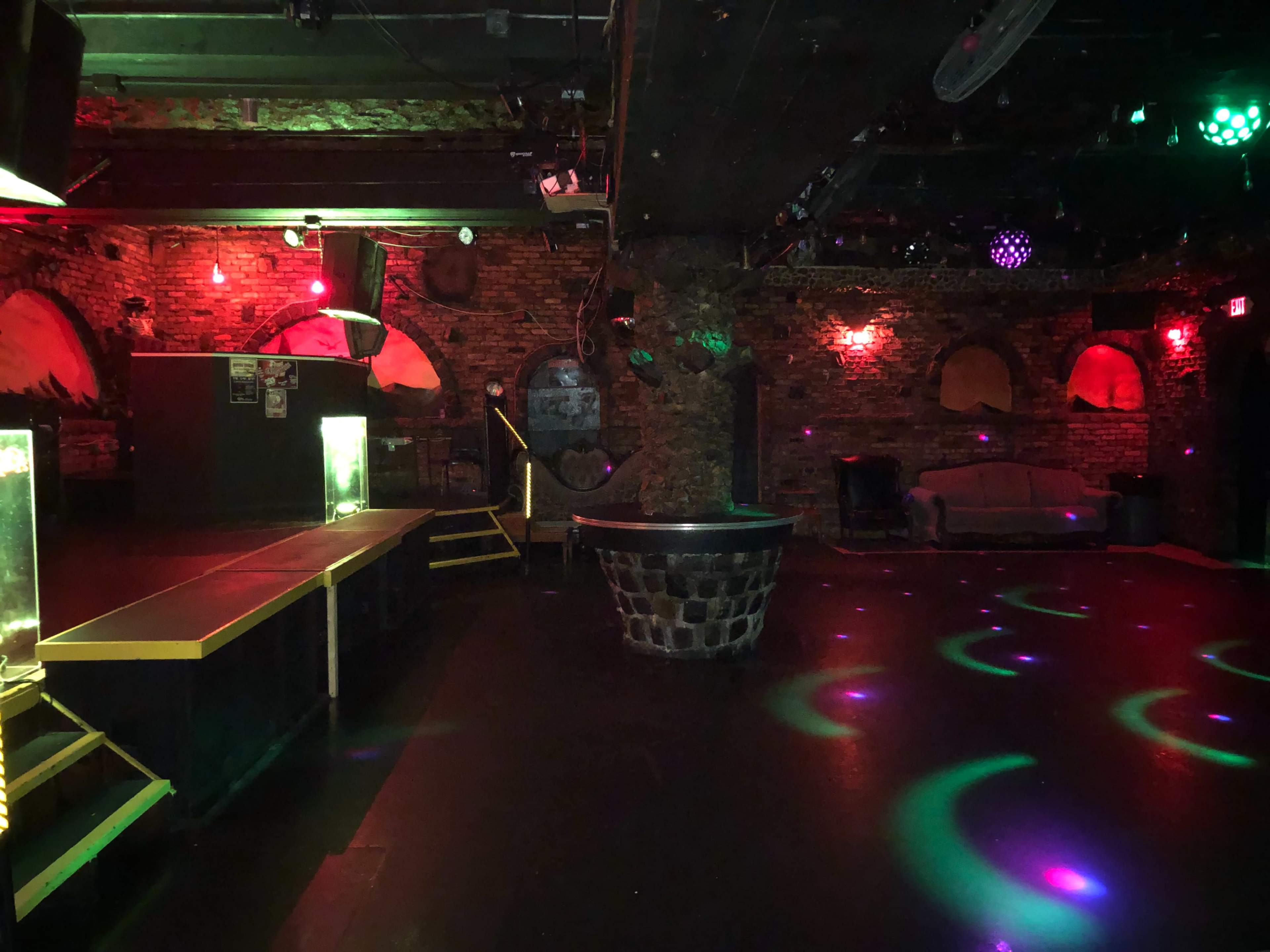A dimly lit nightclub with brick walls, a raised stage, and multi-colored lights illuminating the floor.