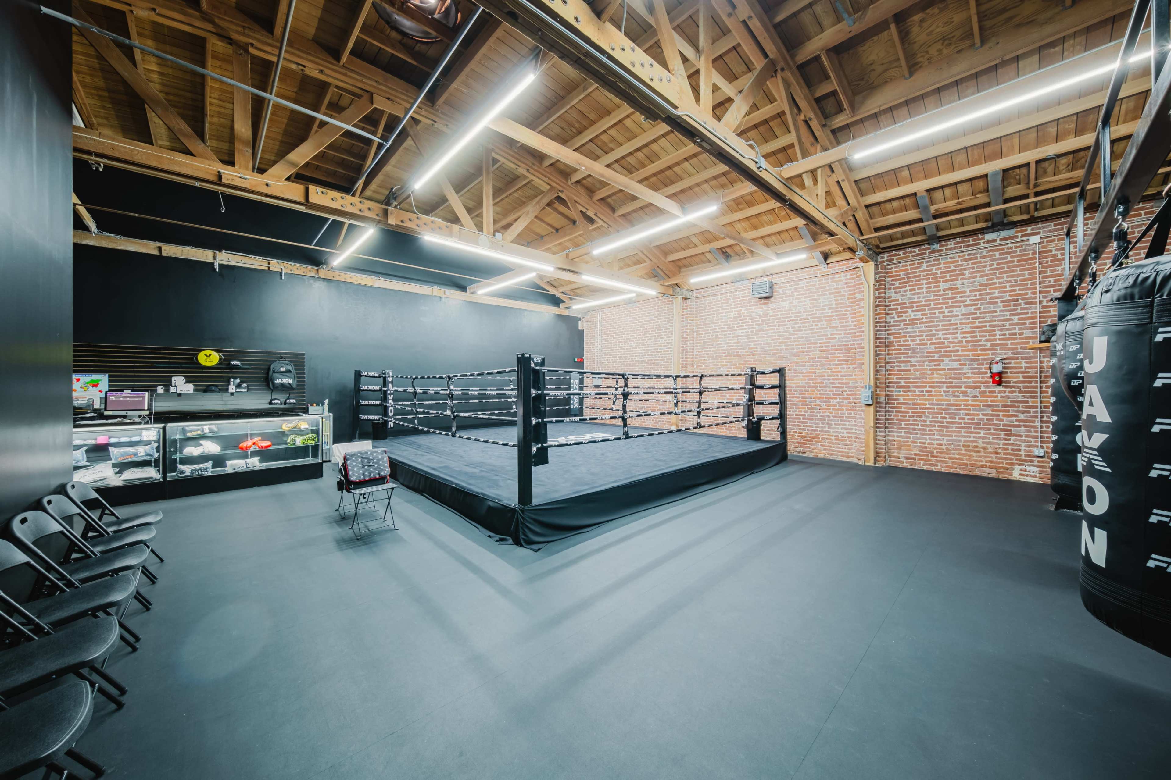 The image shows a boxing gym with a ring in the center, surrounded by black mats and exposed brick walls, with chairs set up along one side.