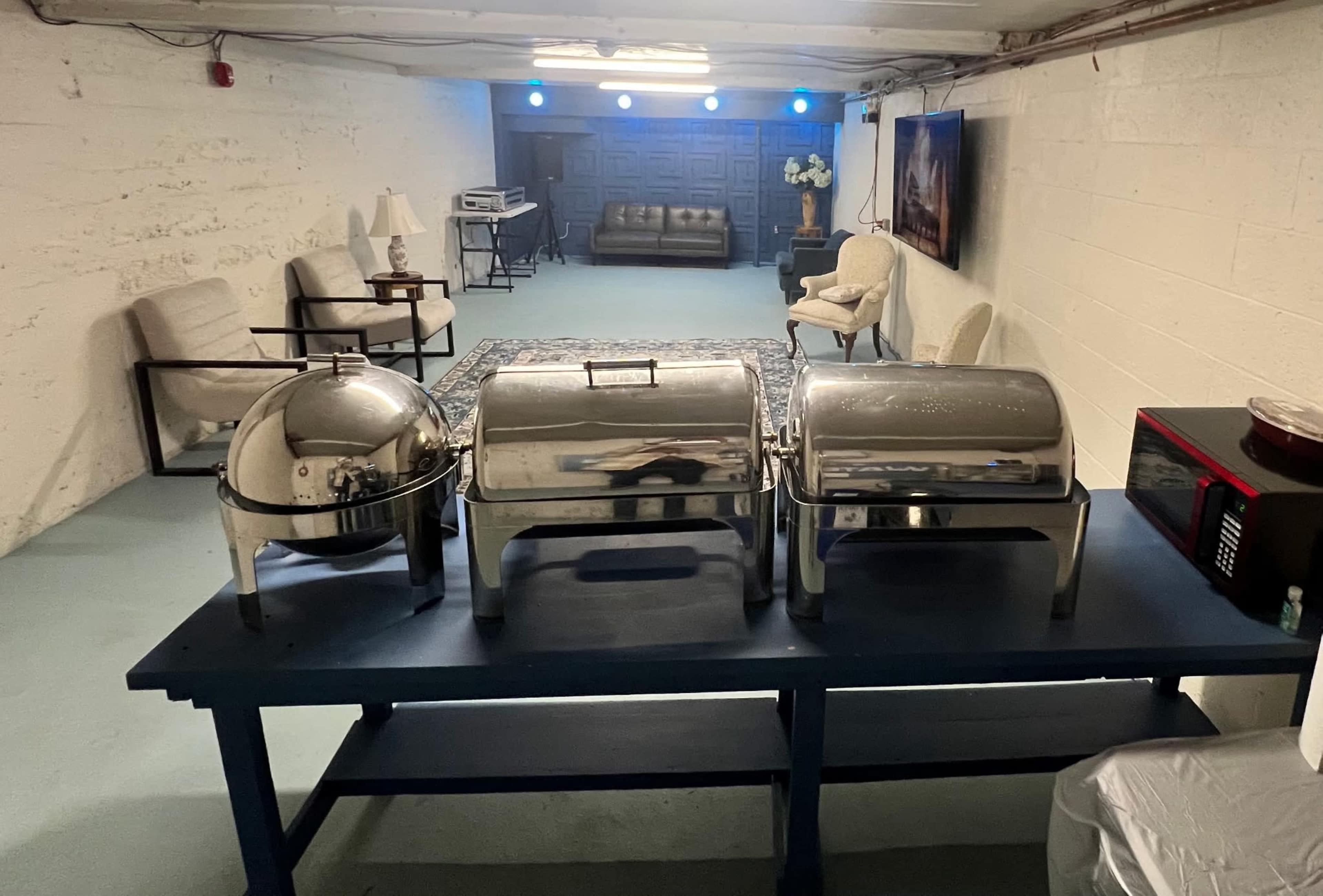 A table with three stainless steel chafing dishes is positioned in a basement room that features a couch and chairs against a white wall.