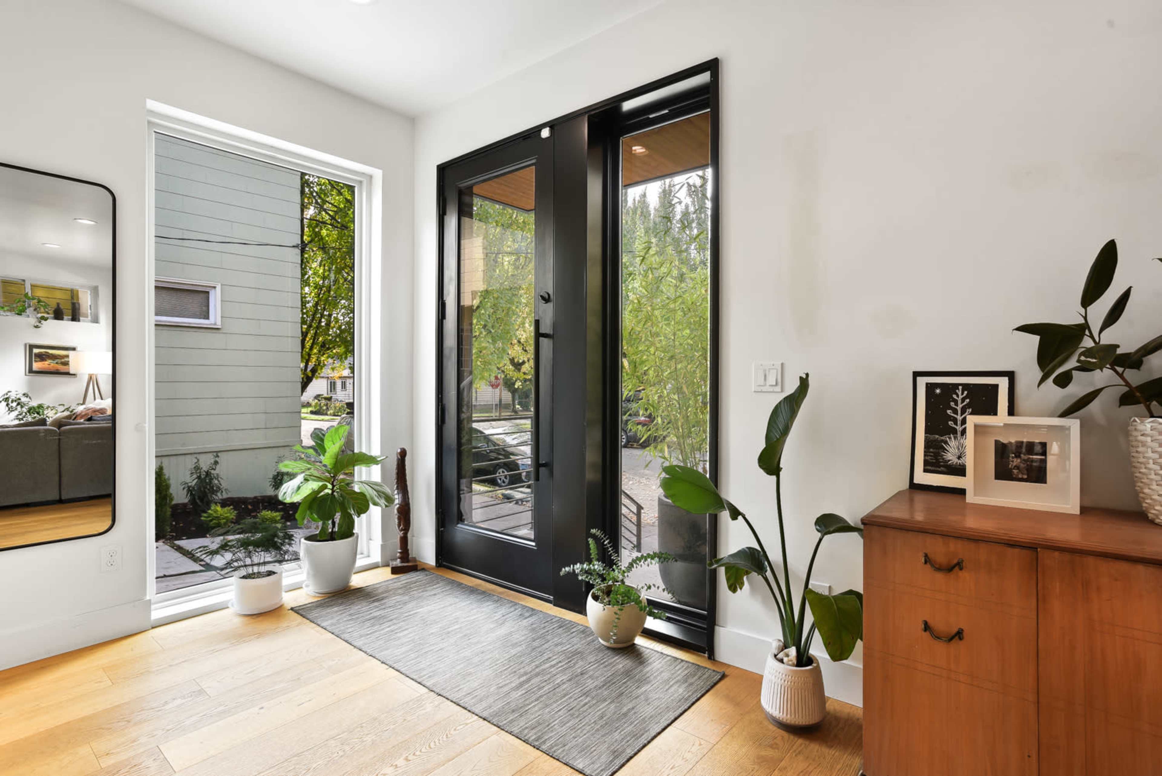 The entryway features a pair of black glass doors, a rug, potted plants, and a wooden cabinet with framed pictures.