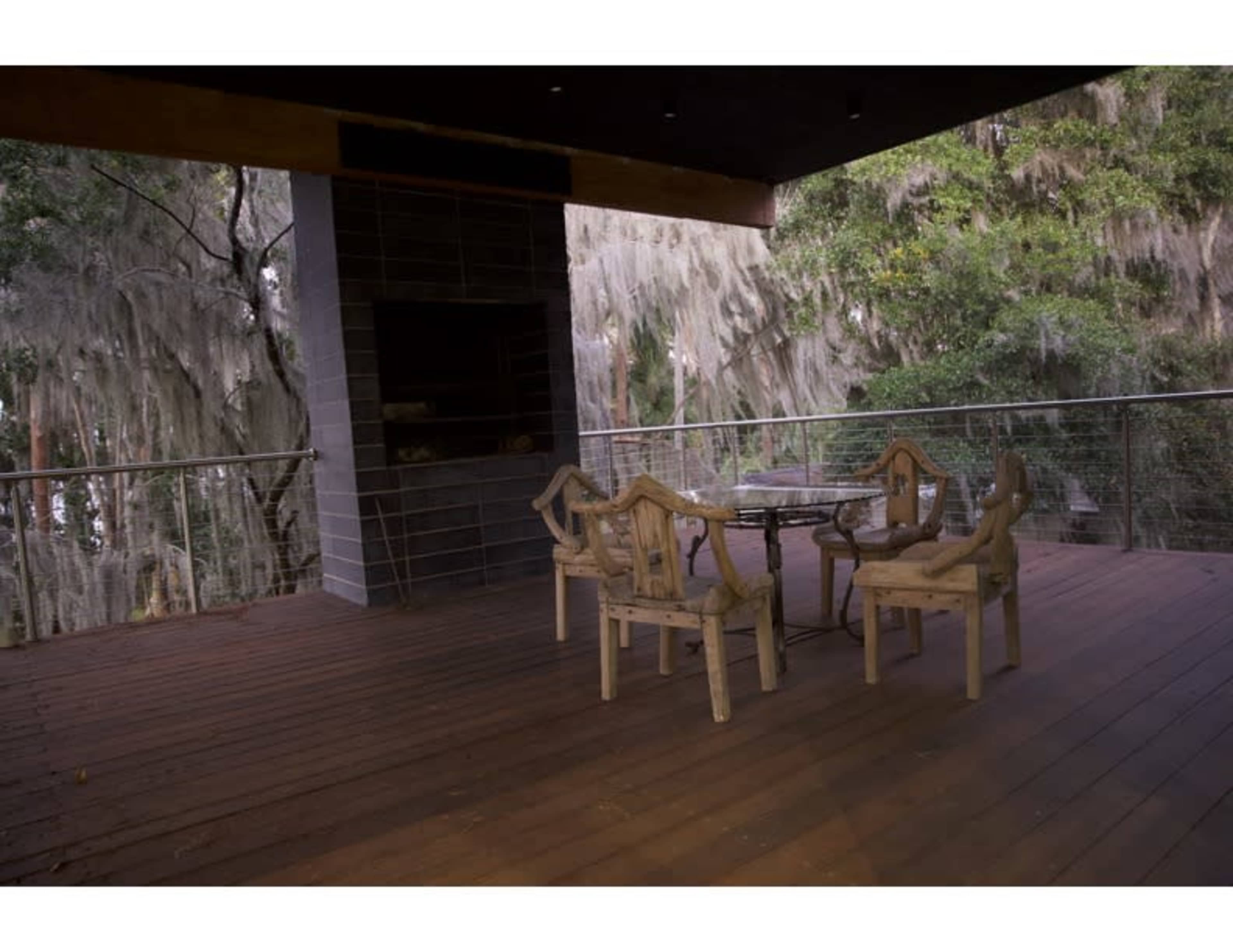 A wooden deck with a circular glass table and six wooden chairs overlooks a wooded area covered in Spanish moss.