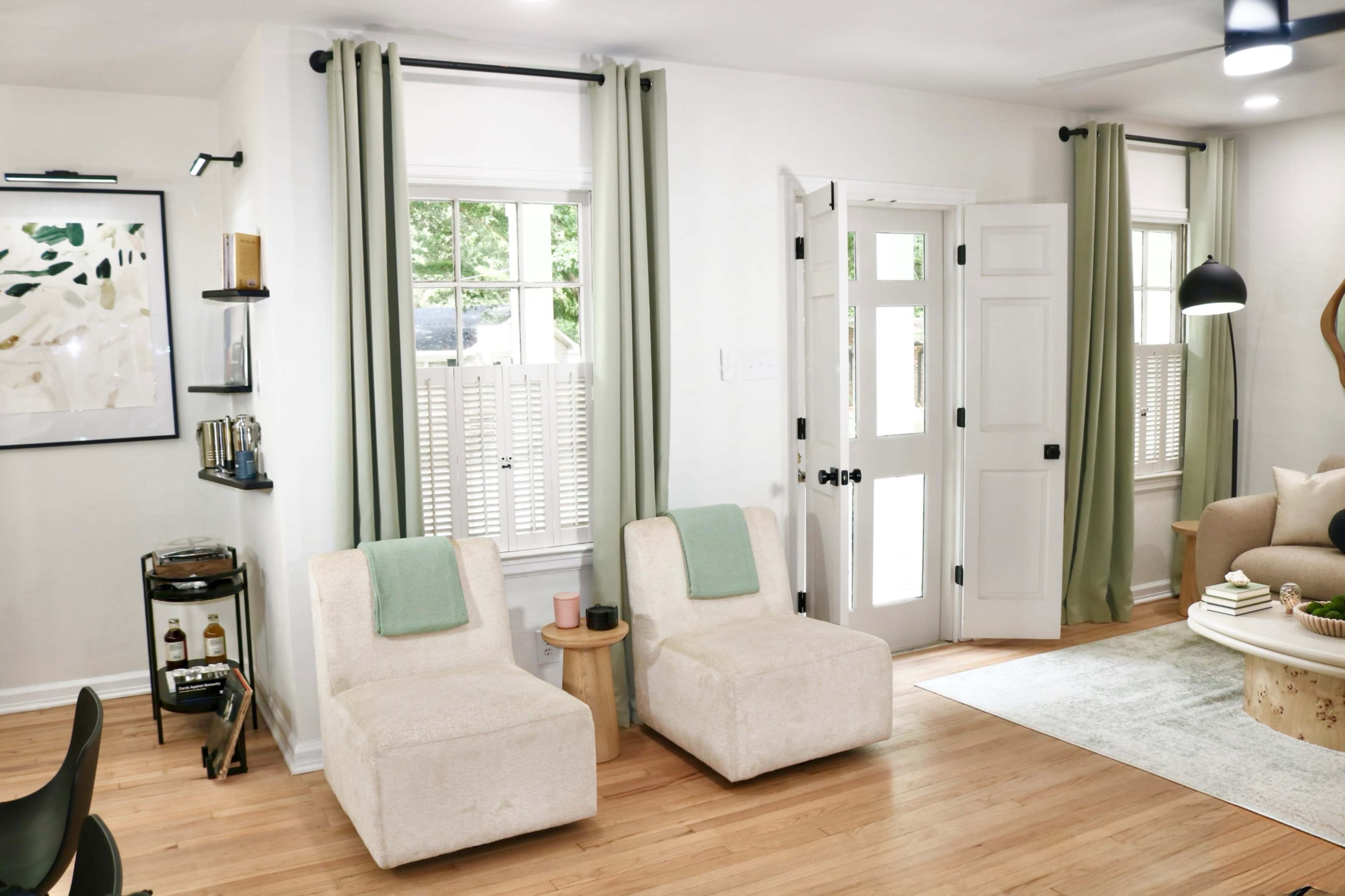 The image shows a bright living room featuring two cream-colored armchairs, a small side table, and large windows with light green curtains.