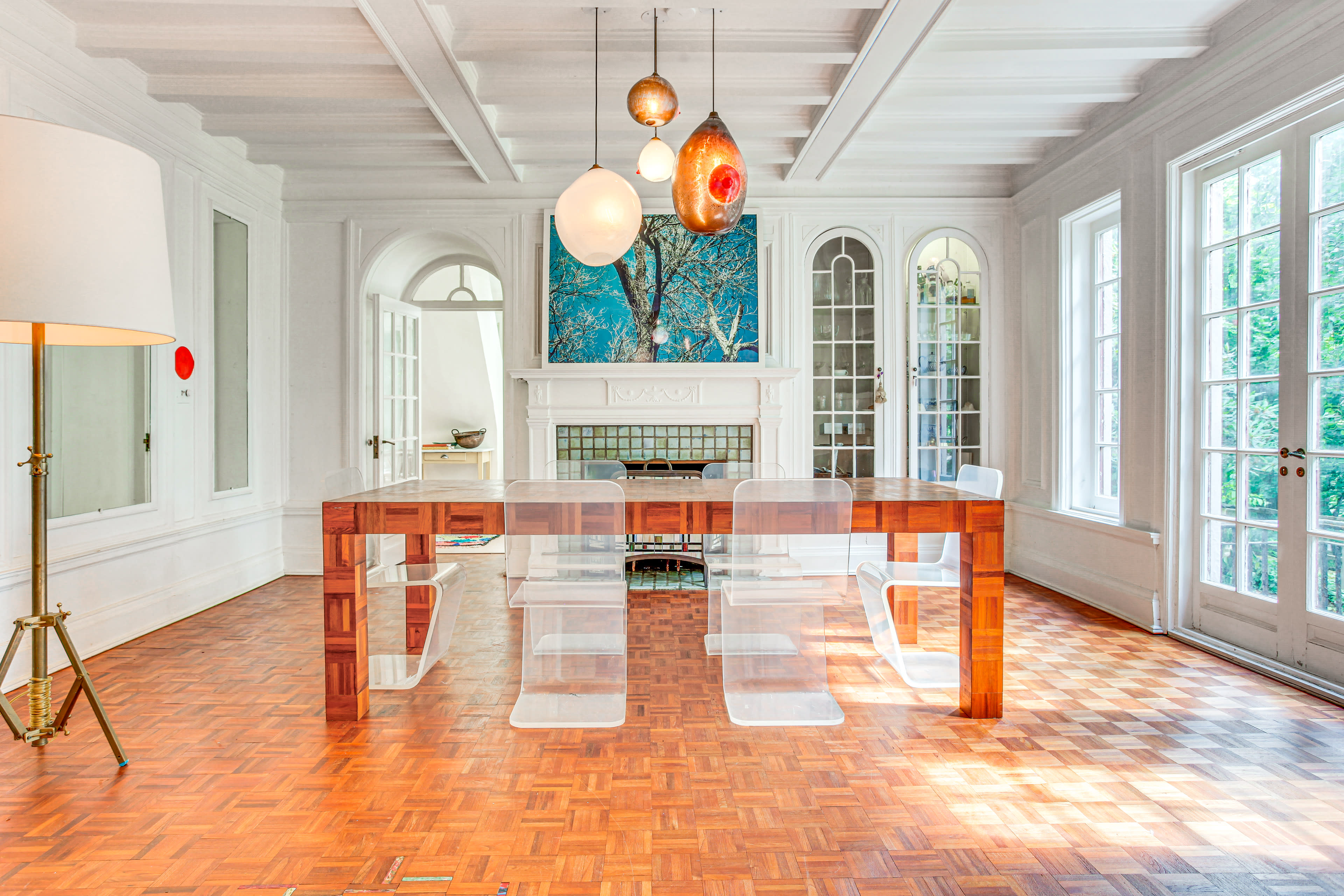 A modern dining room features a wooden table surrounded by clear acrylic chairs, with large windows and contemporary light fixtures above.