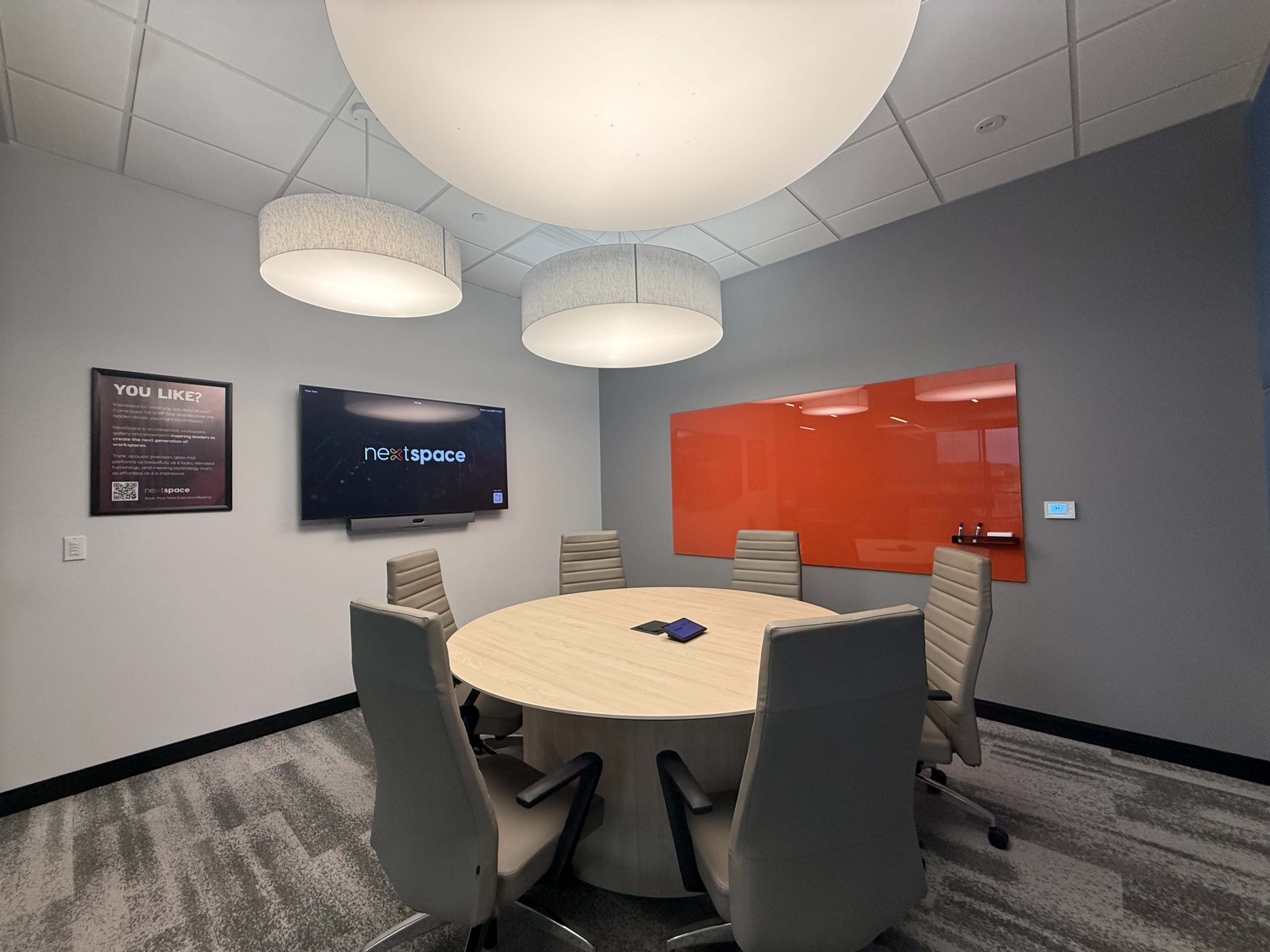 The image shows a modern conference room with a round wooden table, six high-backed chairs, a large screen on the wall, and a colorful orange whiteboard.