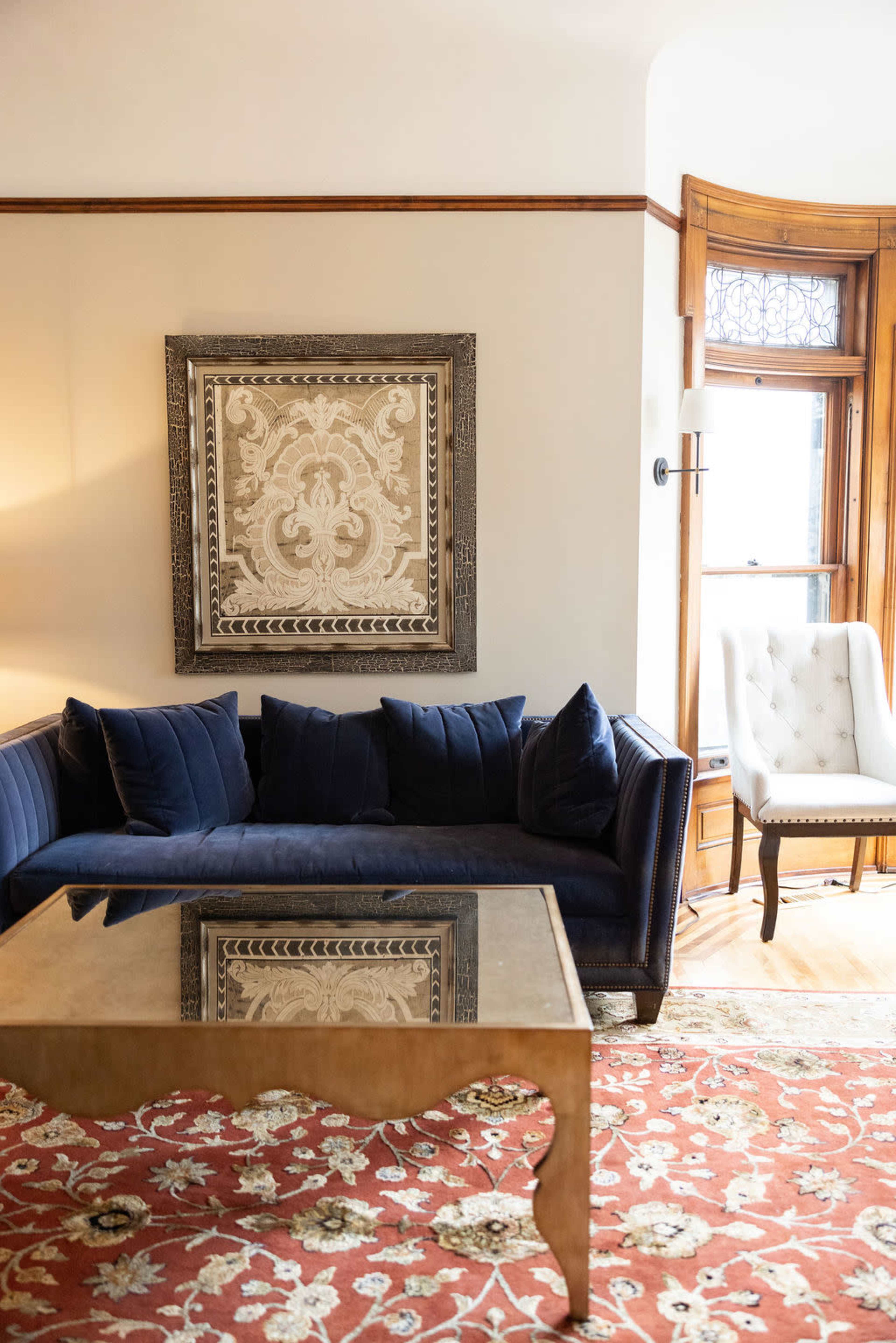 The image shows a living room featuring a navy blue sofa with several cushions, a glass-top coffee table, an intricately framed wall art piece, and a light-colored armchair beside a window.