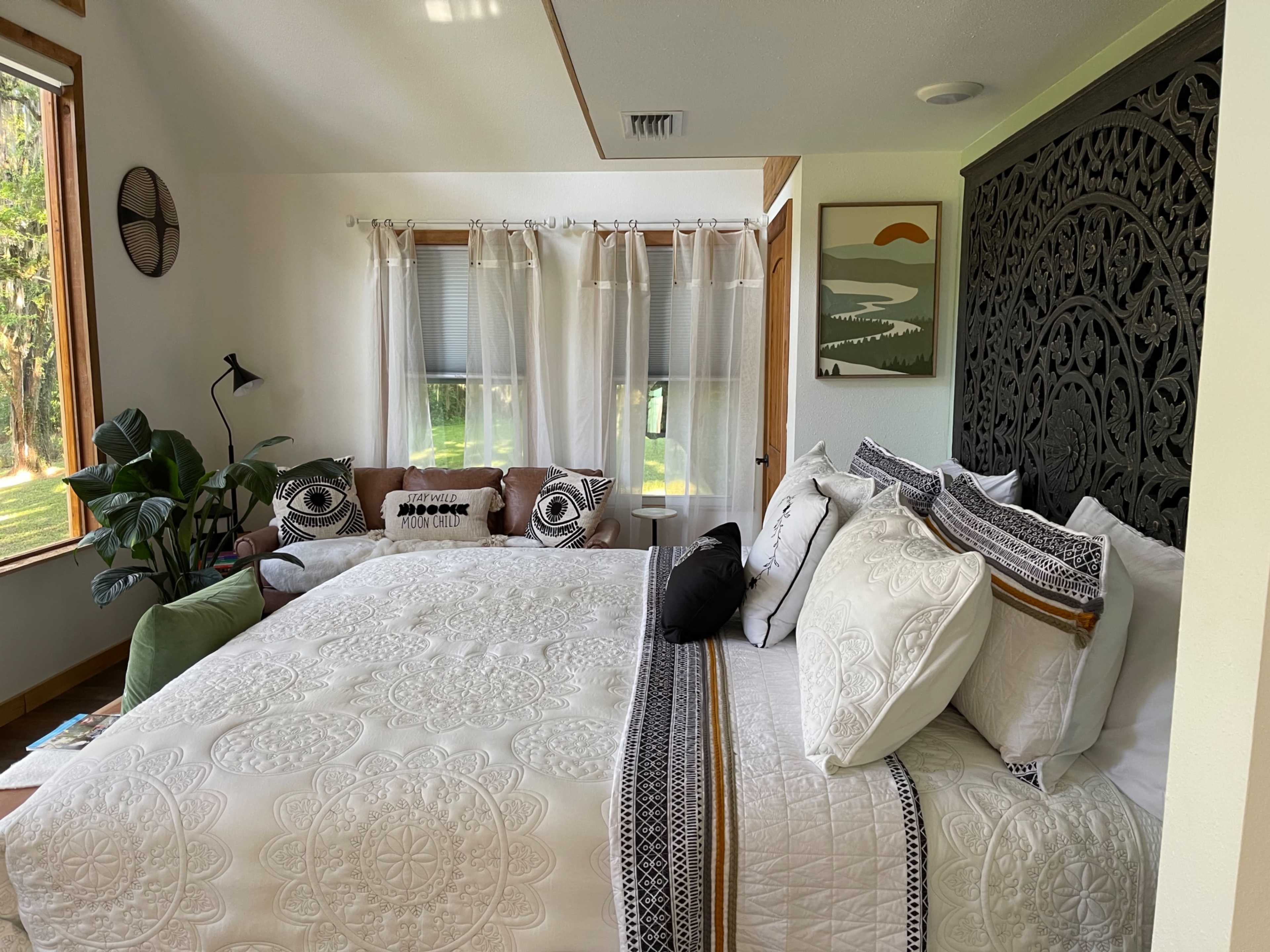 A well-decorated bedroom features a large bed with intricate bedding, a cozy seating area with cushions, and large windows letting in natural light.